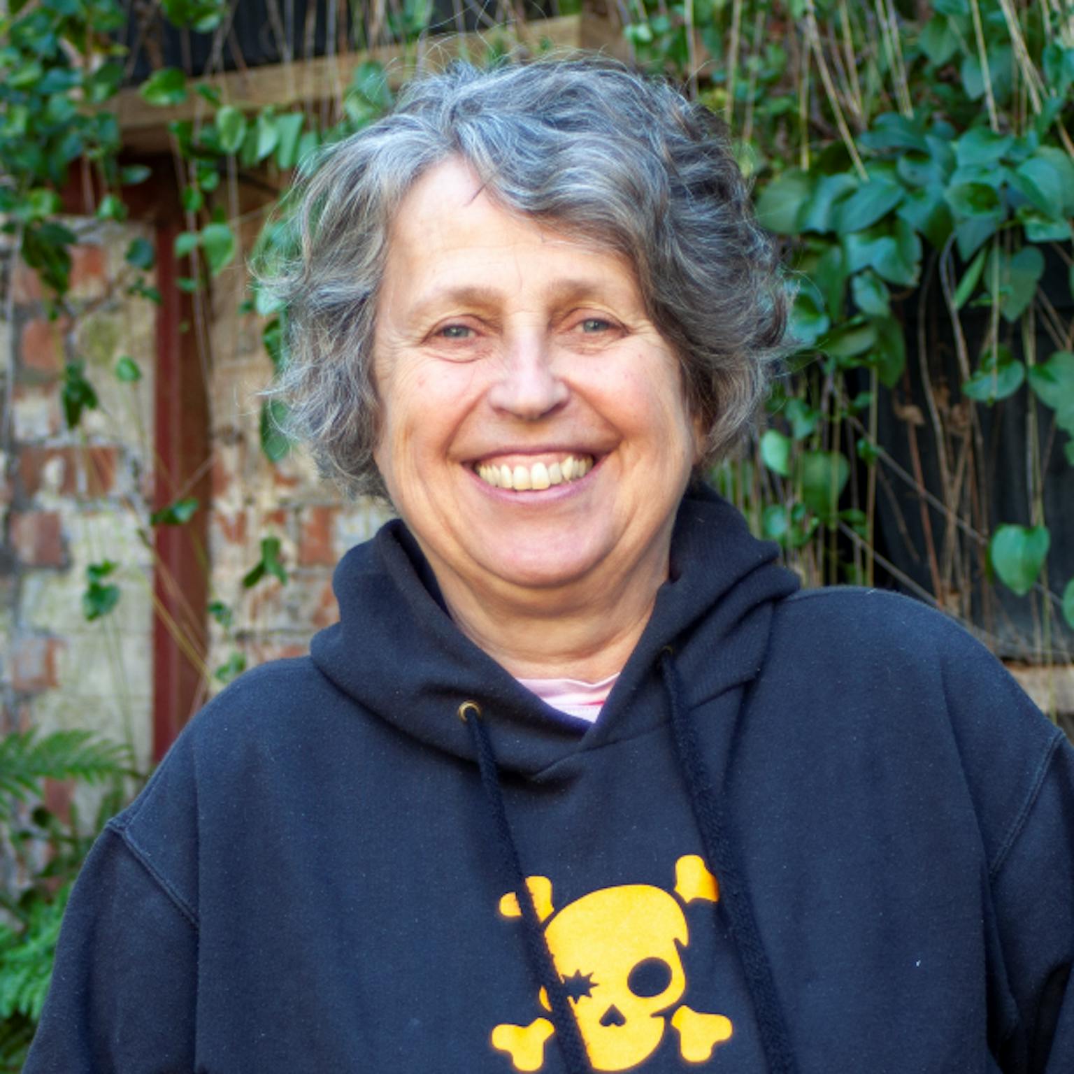 A person standing in front of greenery, she is wearing a black hoodie with a walk the plank logo in the centre. She is smiling with her hands in the hoodie pocket