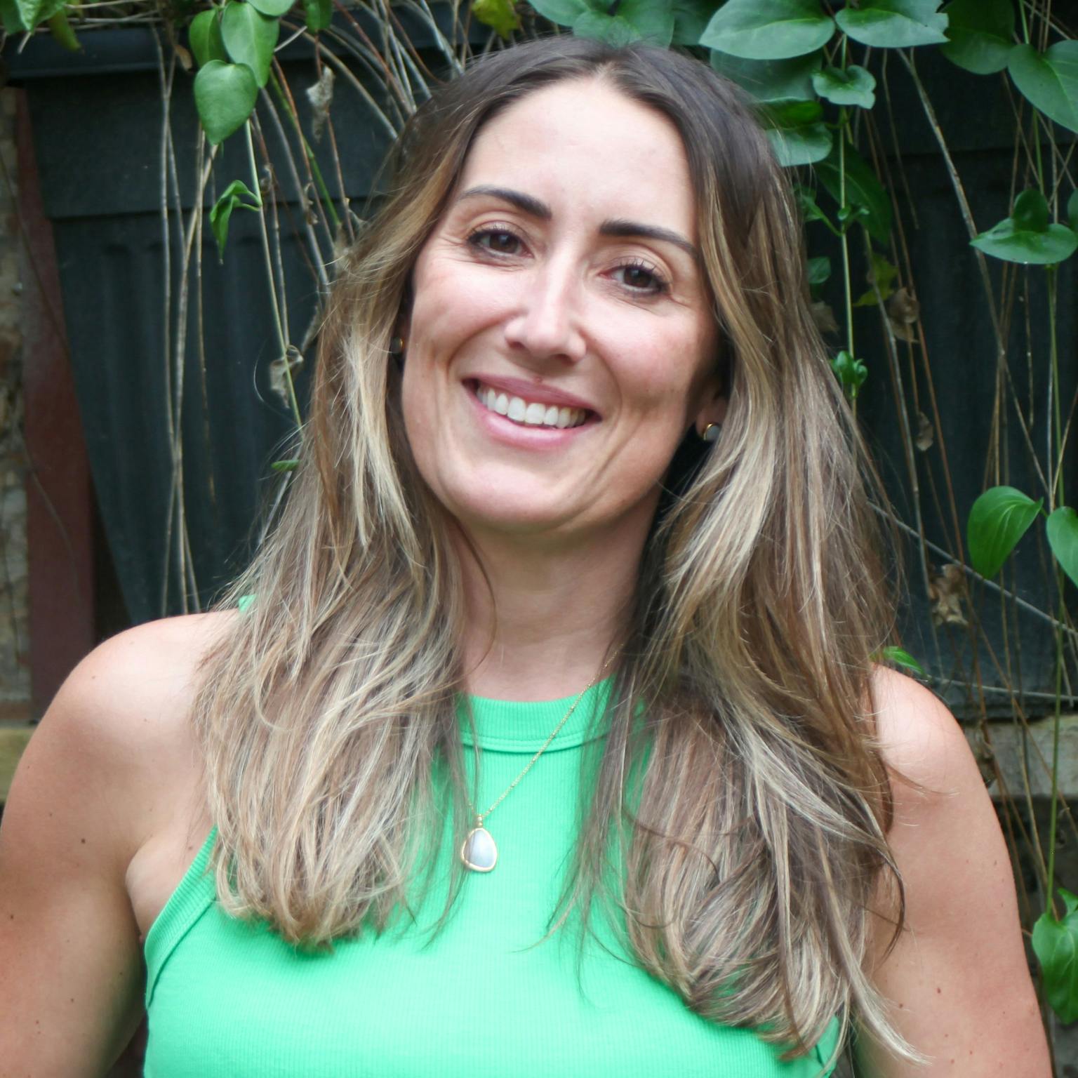 A person standing in front of greenery, she is wearing a green vest top tucked into a pair of jeans. She is smiling with her hands in her pockets