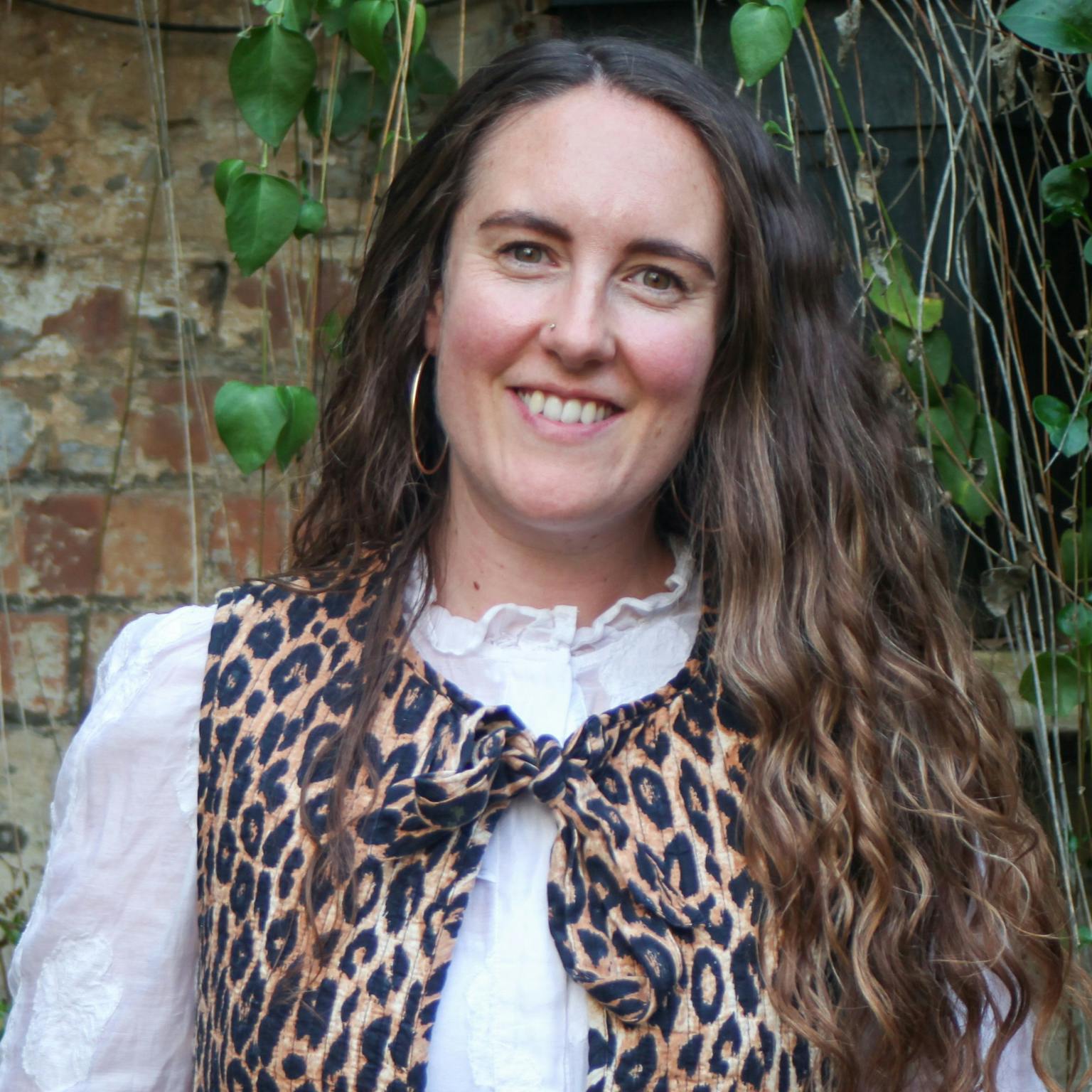 A person standing in front of greenery, she is wearing a leopard print vest over a white blouse, she has long wavy brown hair. She is smiling with her hands in her pockets