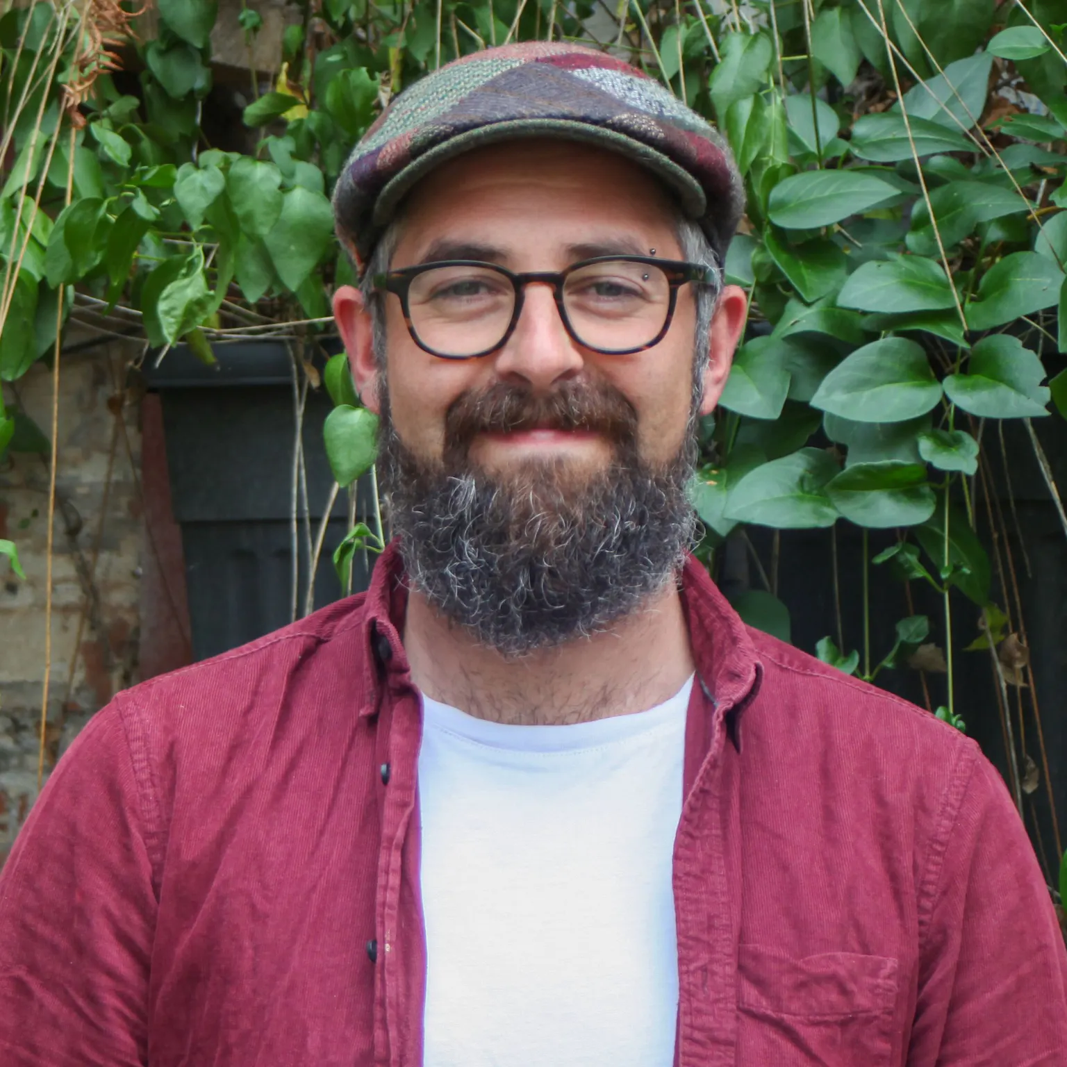 A person standing in front of greenery, he is wearing a red shirt over a white undershirt, he has a patchwork flat-cap and a pair of rounded glasses, he has a dark beard. He is smiling with his arms by his sides.