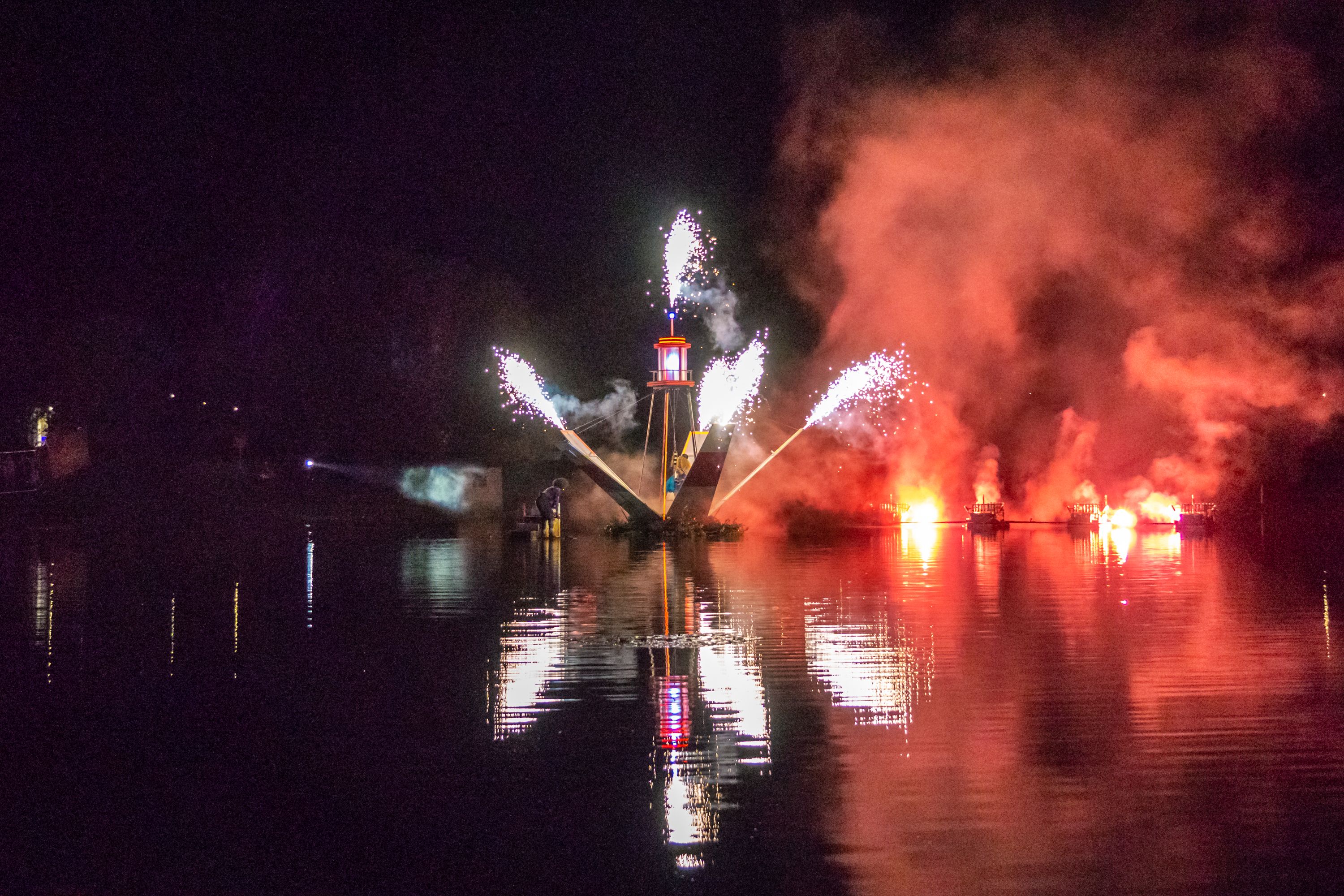 A lighthouse-like structure floating on a lake spraying sparks in multiple directions at night