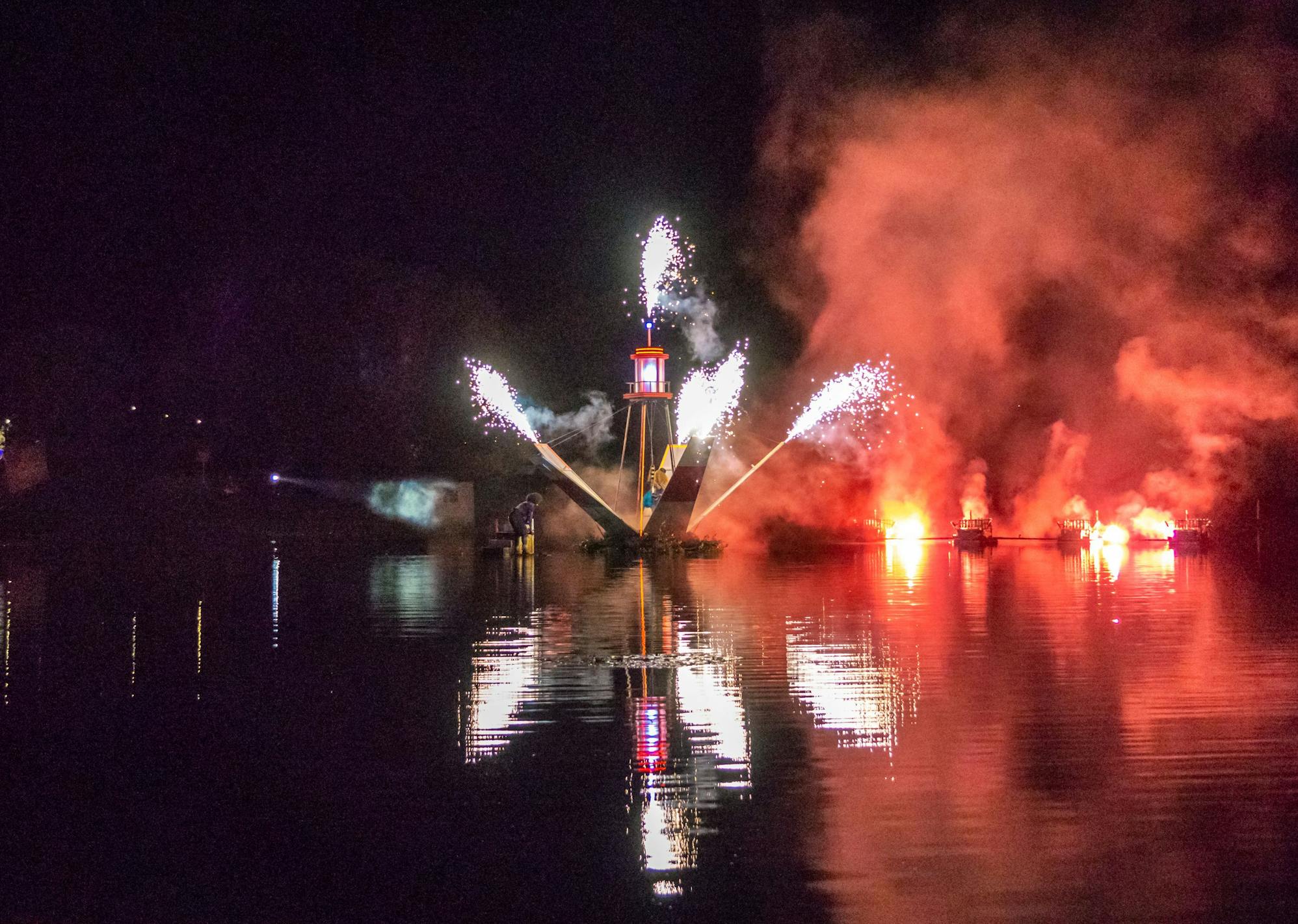 A lighthouse-like structure floating on a lake spraying sparks in multiple directions at night