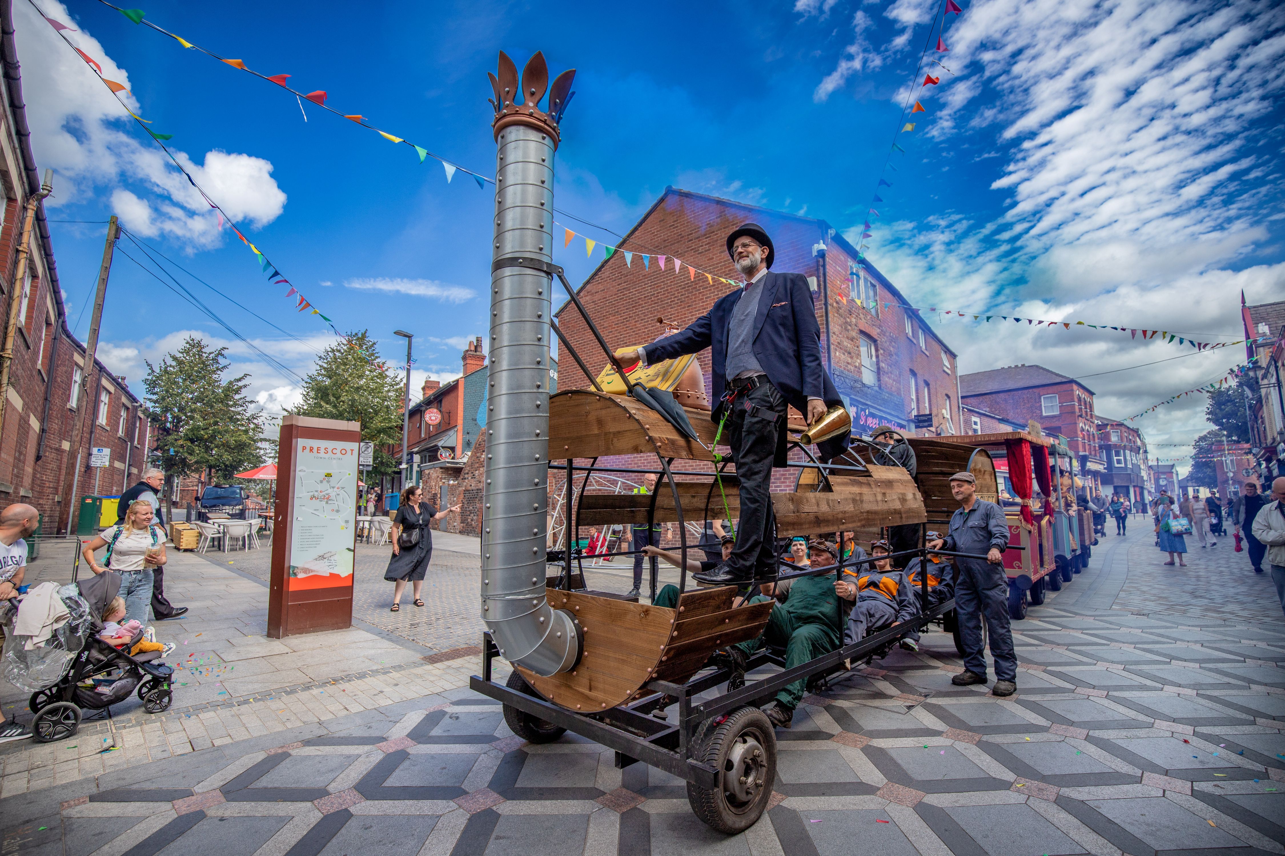 A wooden train on a high street, a performer in a suit and top hat is hanging off the entrance, against a bright blue sky