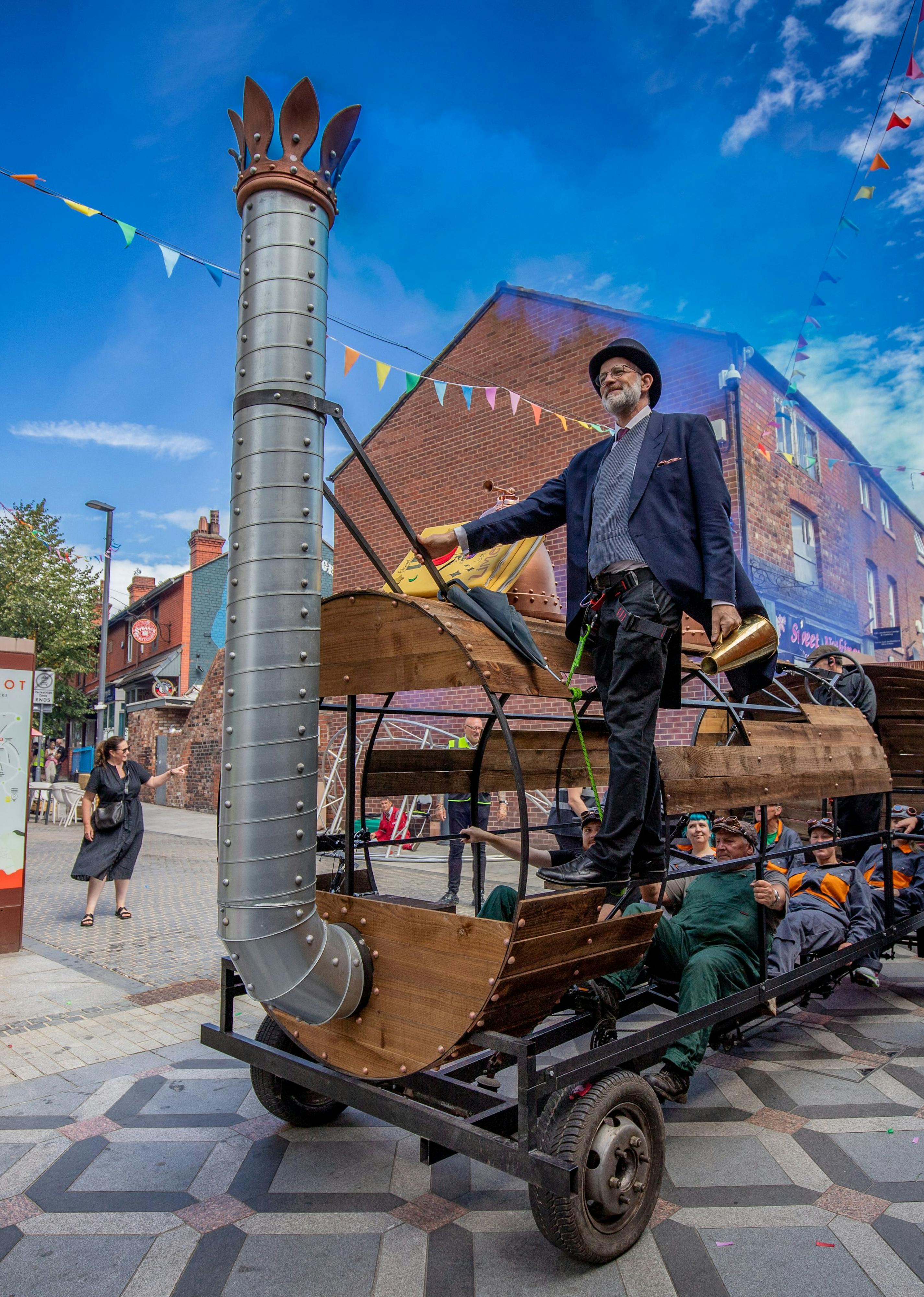 A wooden train on a high street, a performer in a suit and top hat is hanging off the entrance, against a bright blue sky