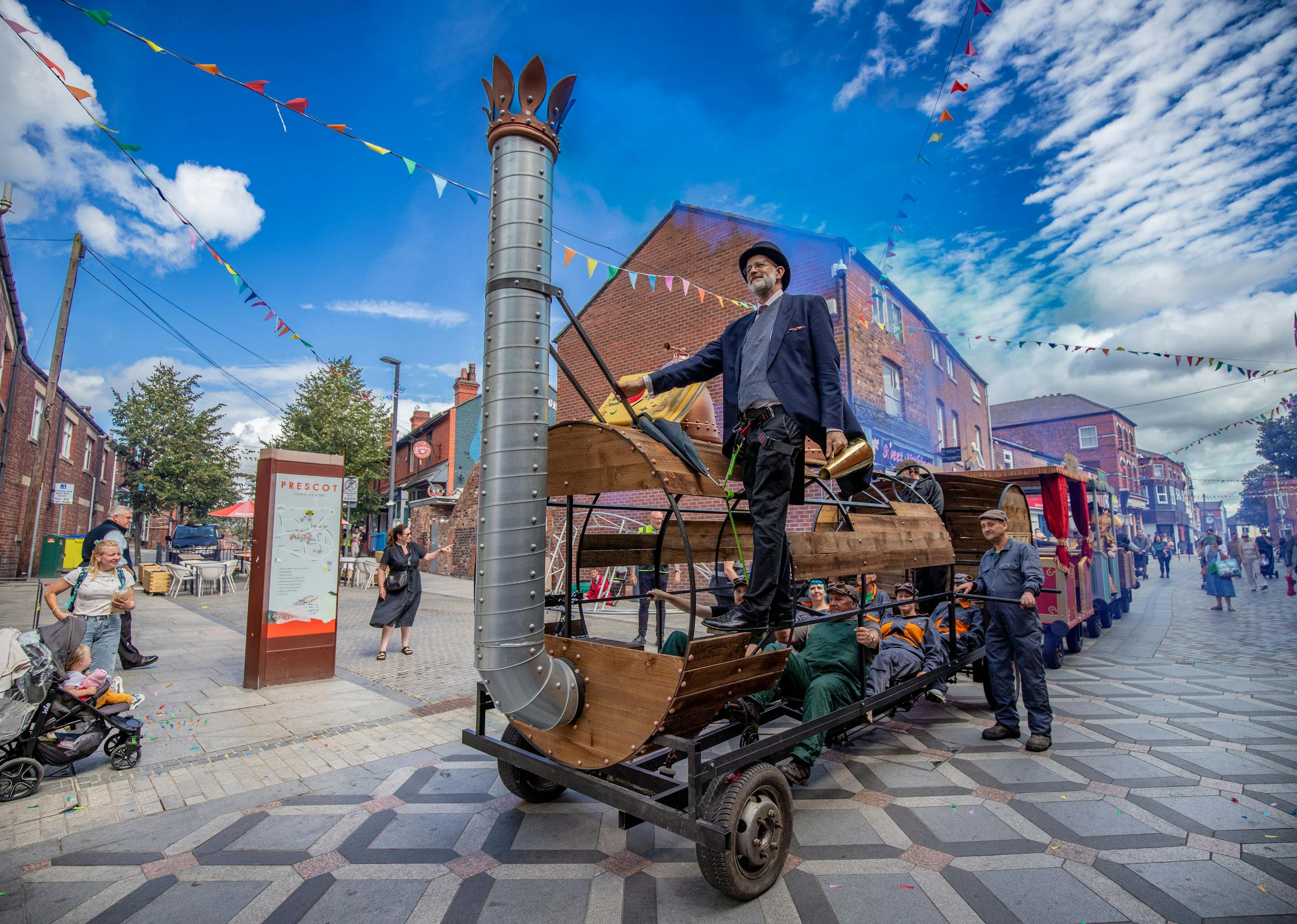 A wooden train on a high street, a performer in a suit and top hat is hanging off the entrance, against a bright blue sky