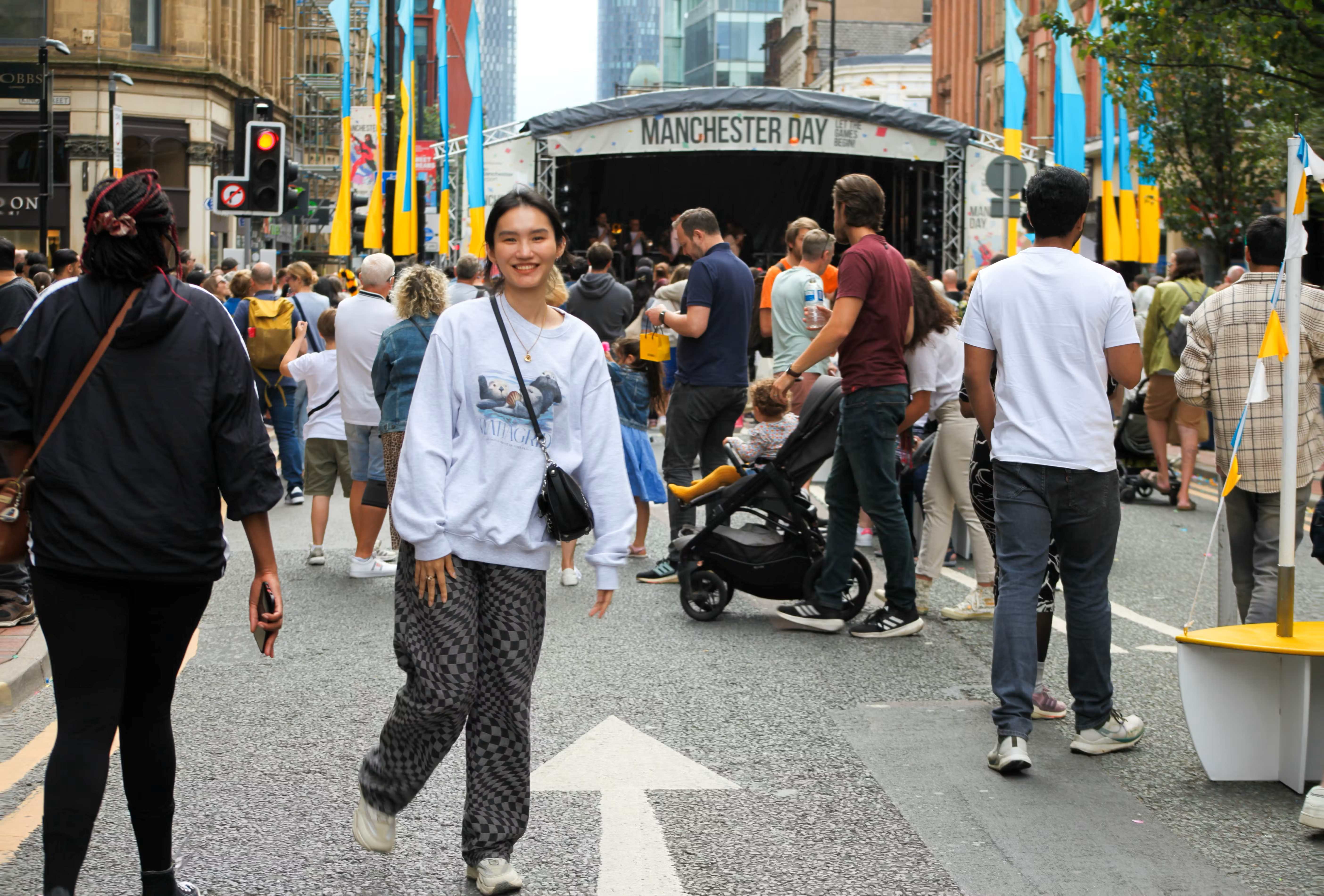 A person in a grey jumper and black and grey trousers posing for a photo in front of a Manchester Day stage, surrounded by people.