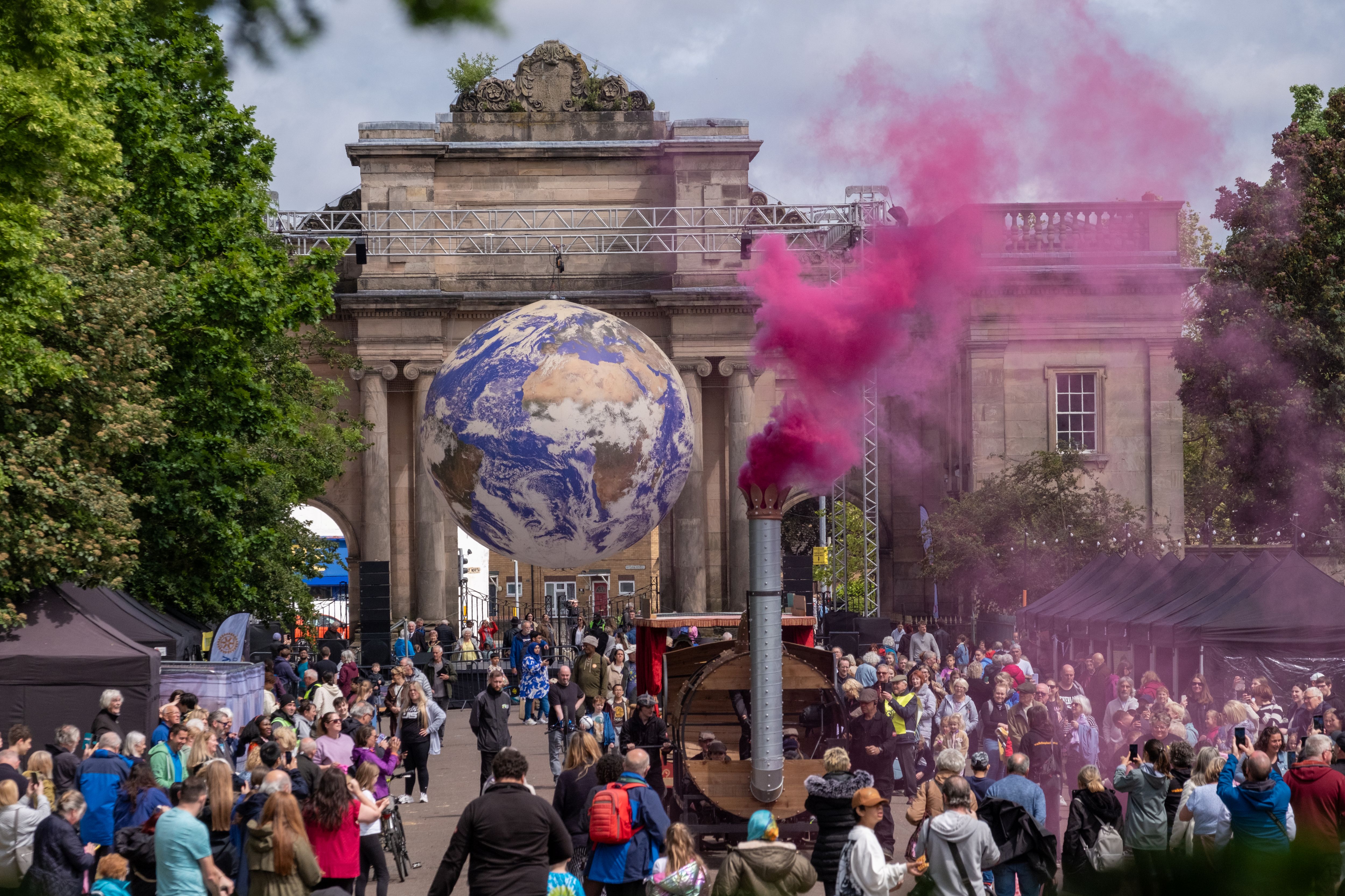 A train float with three carriages carrying pantomime horses in park as crowd look on with pink smoke.