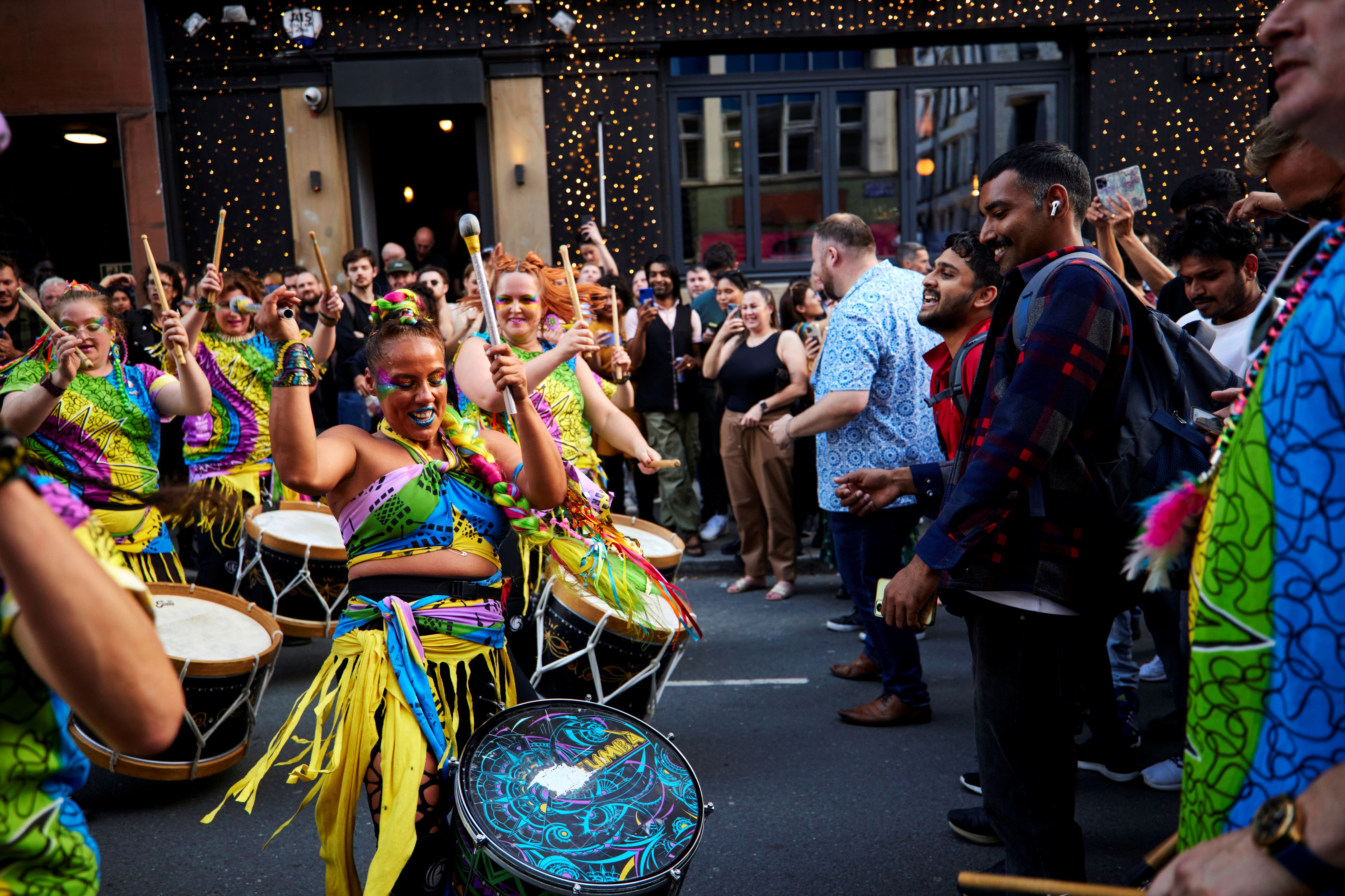 A procession of people dressed in colourful outfits drumming in front of a crowd of onlookers with their phones out, taking photos