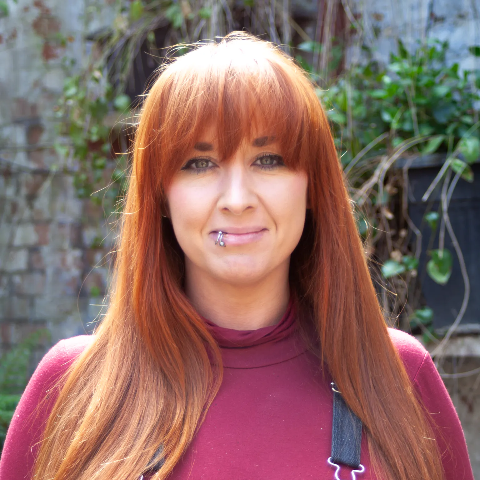A person with long straight red hair standing in front of greenery. She is wearing a burgundy top under dungarees and has a piercing on her lip
