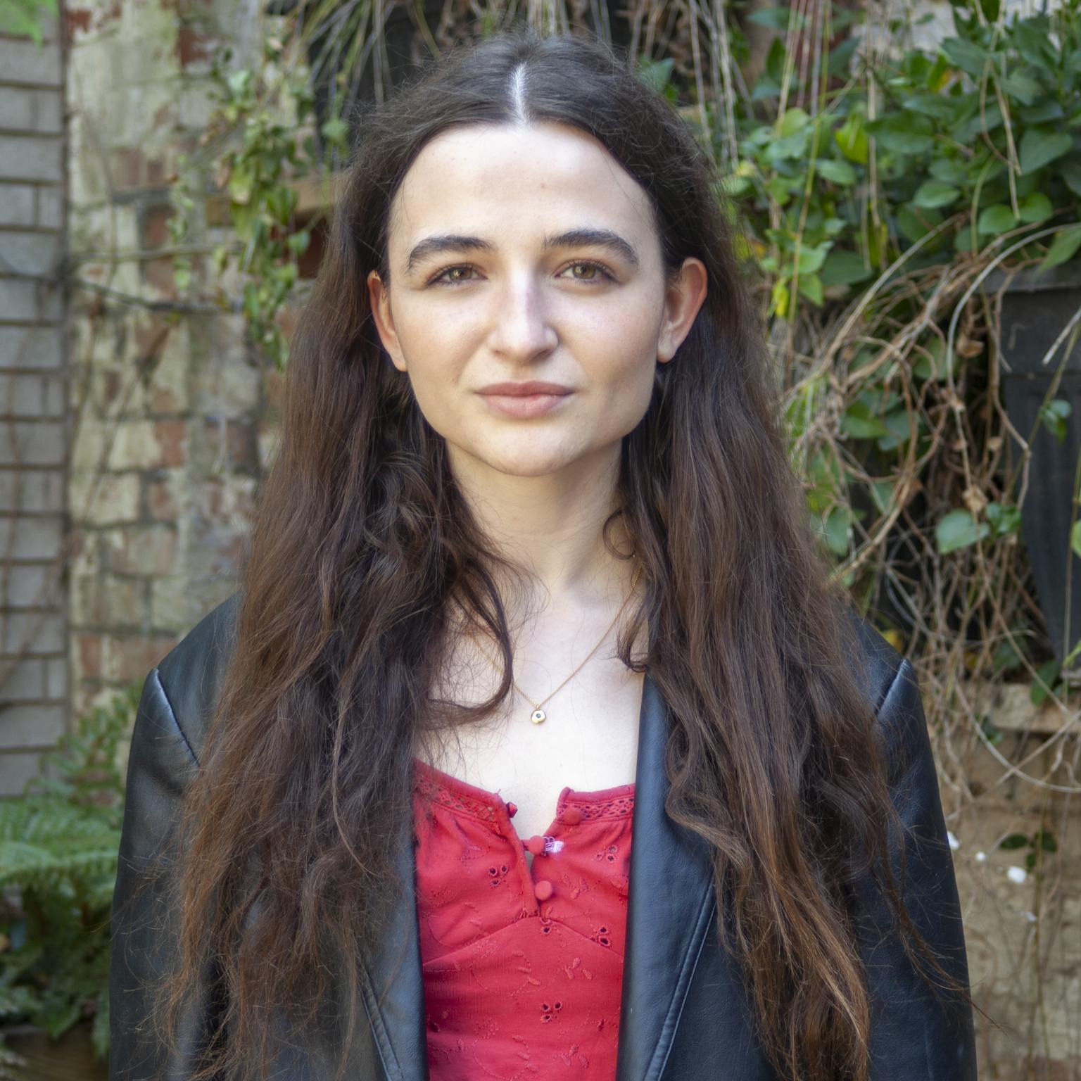 A person with long dark hair standing in front of greenery. They are wearing a red top and a black leather blazer