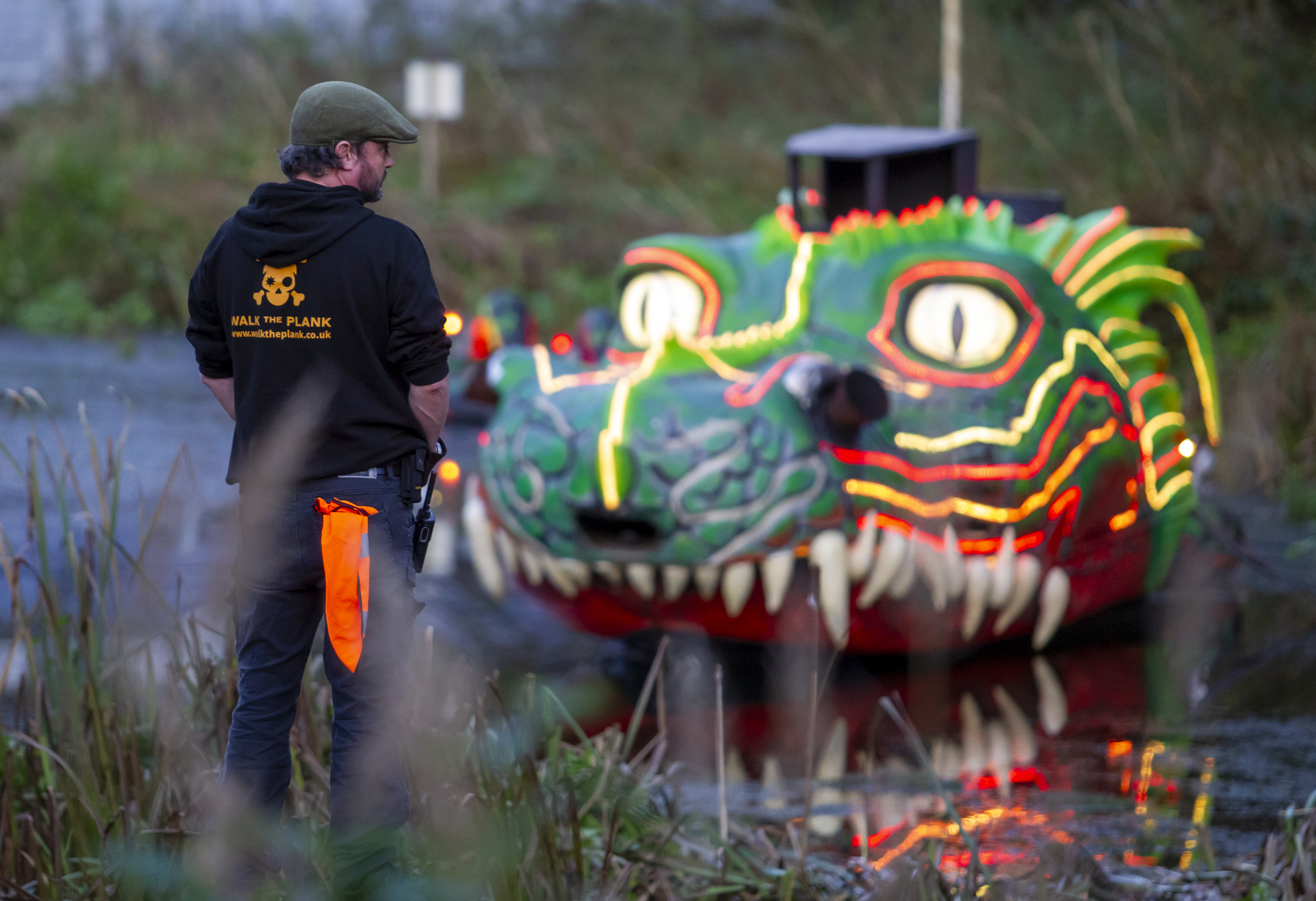A person in a green cap and black hoodie with a orange skull and cross bones on it looking at a body of water with a dragon's head artwork on.