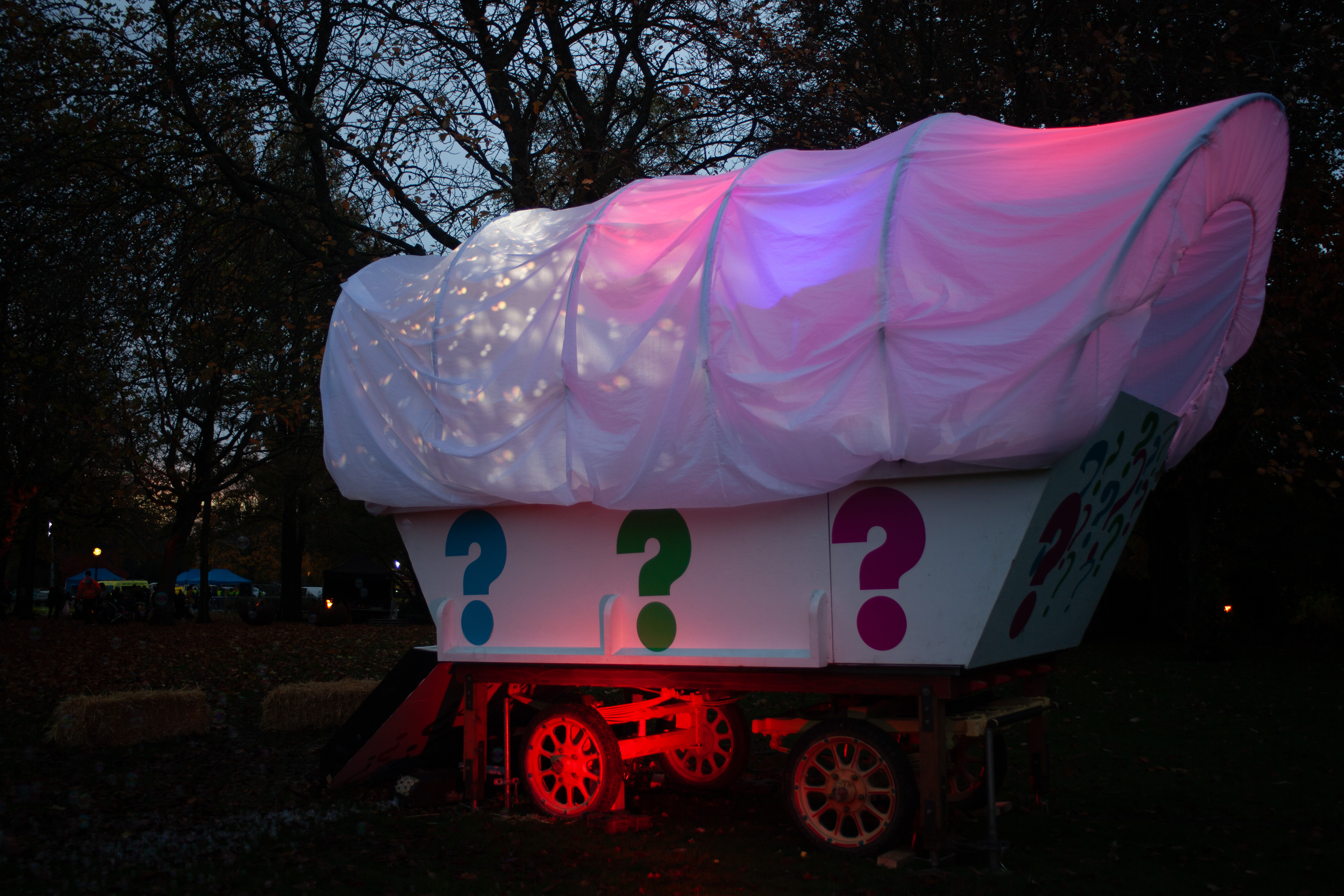 A giant white wagon structure lit up with pink, red and purple lighting, with colourful question marks painted on the side, in a woodland area at night.