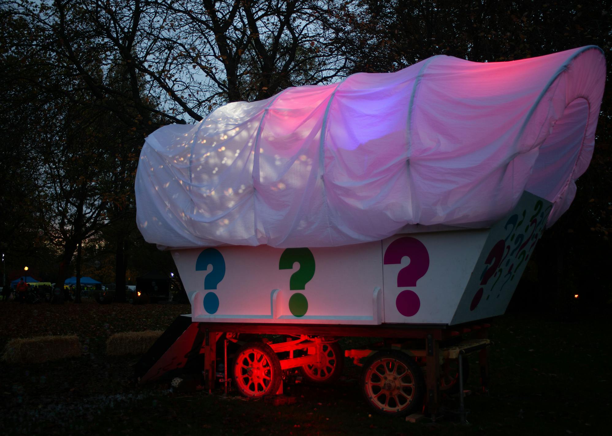 A giant white wagon structure lit up with pink, red and purple lighting, with colourful question marks painted on the side, in a woodland area at night.
