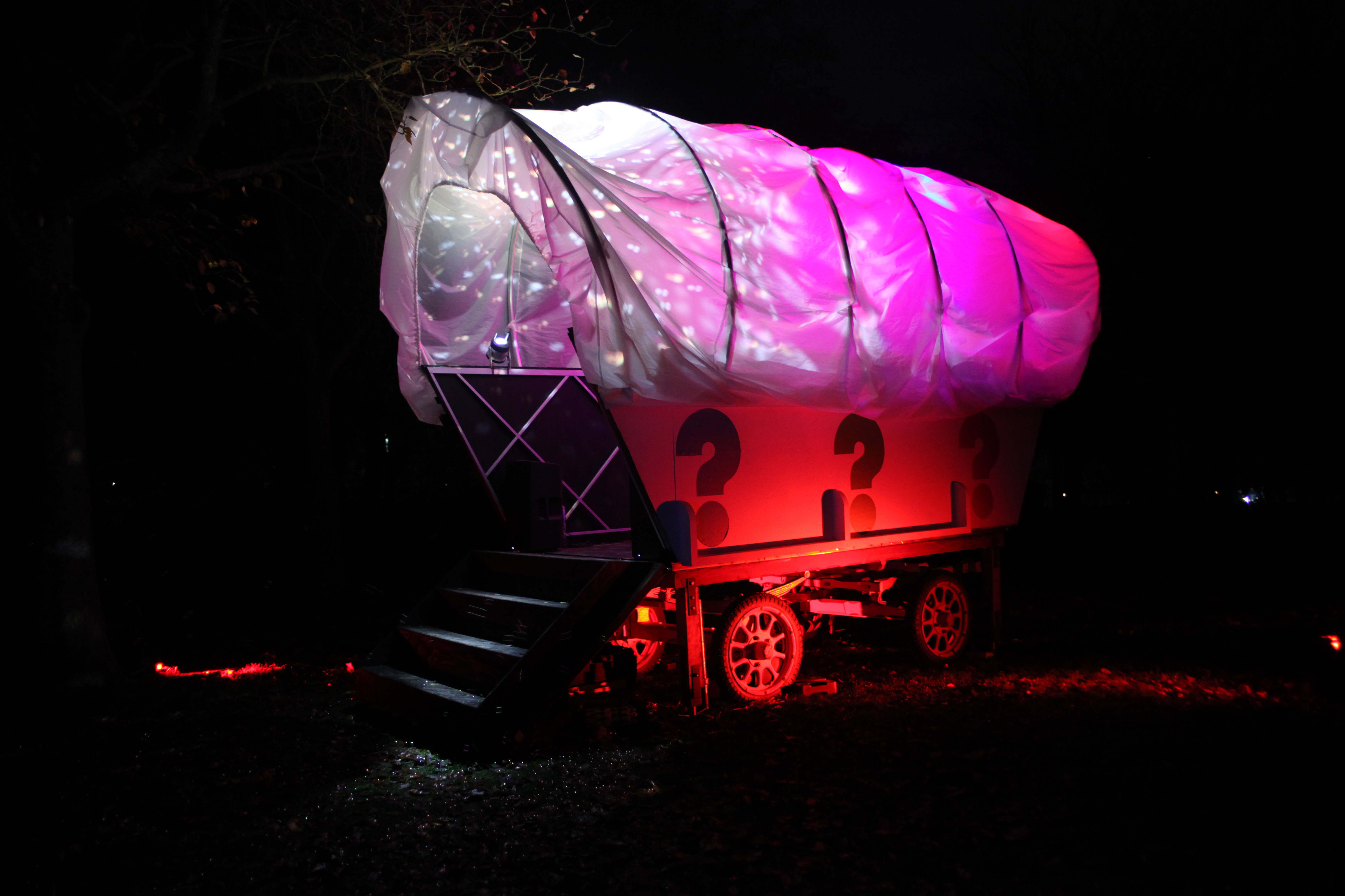 A giant white wagon structure lit up with pink, red and purple lighting, with colourful question marks painted on the side, in a woodland area at night.