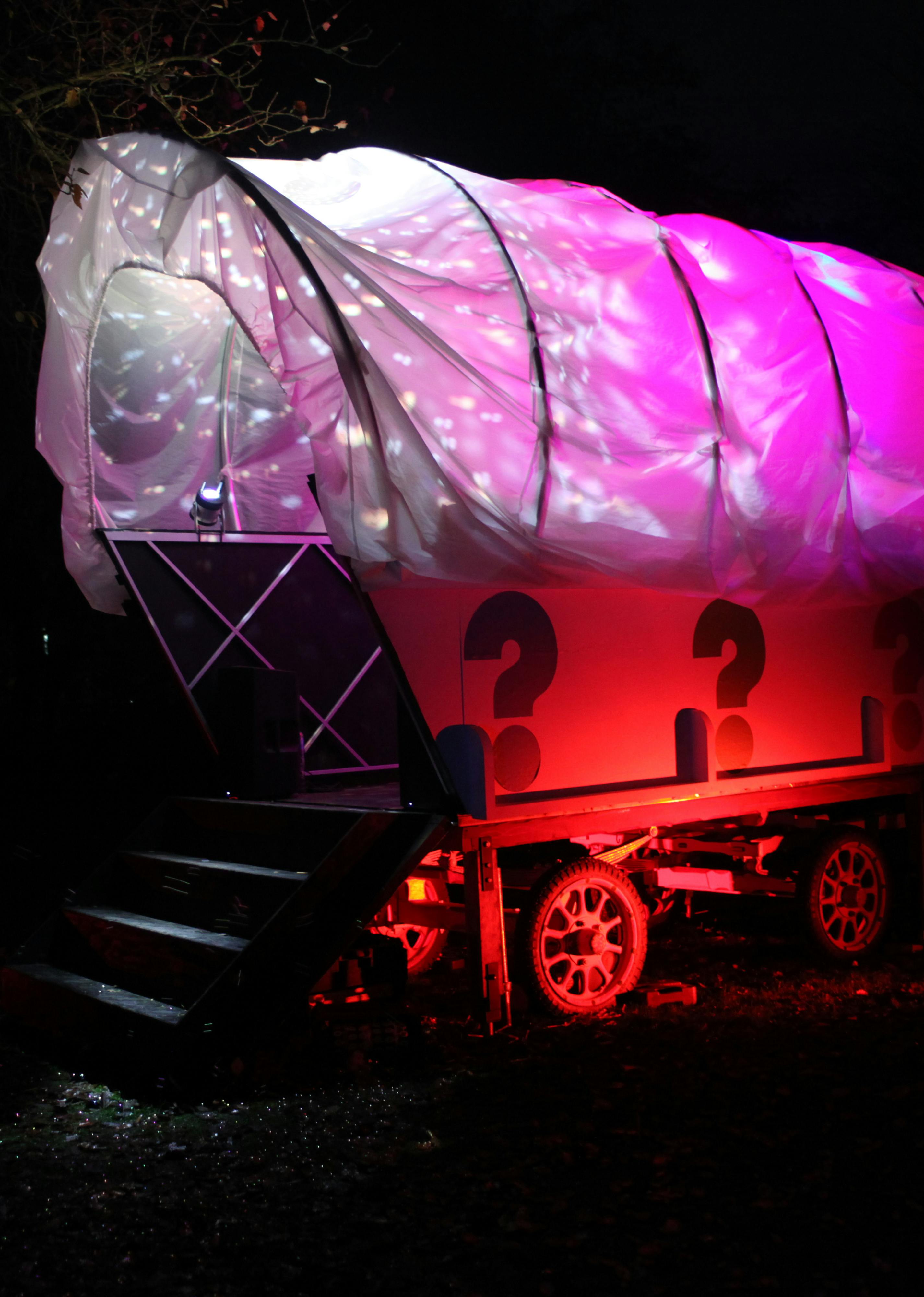 A giant white wagon structure lit up with pink, red and purple lighting, with colourful question marks painted on the side, in a woodland area at night.