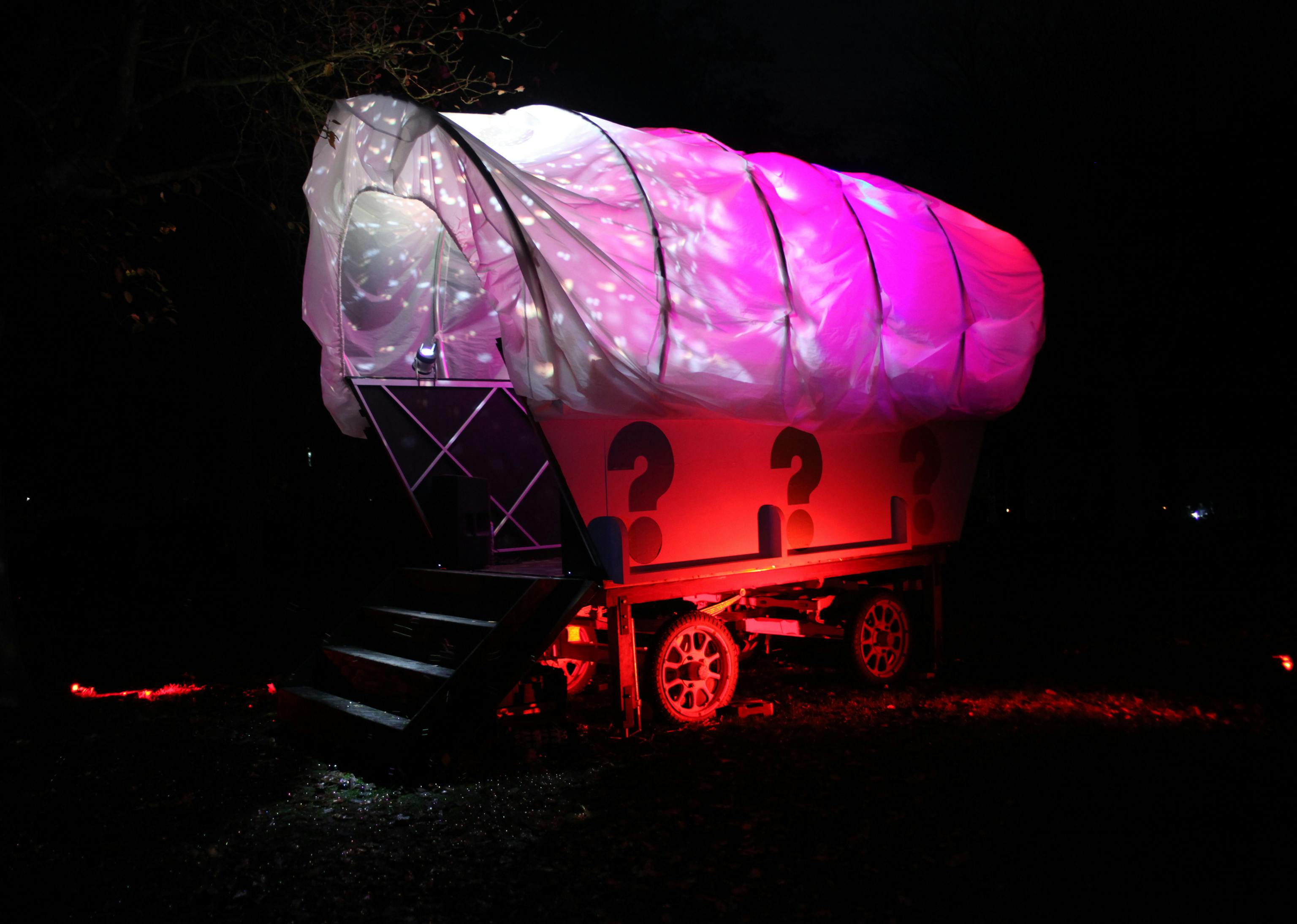 A giant white wagon structure lit up with pink, red and purple lighting, with colourful question marks painted on the side, in a woodland area at night.