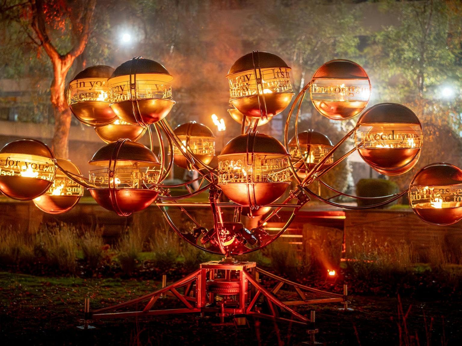 A metal sculpture featuring globes of fire with text about love of them, at night.