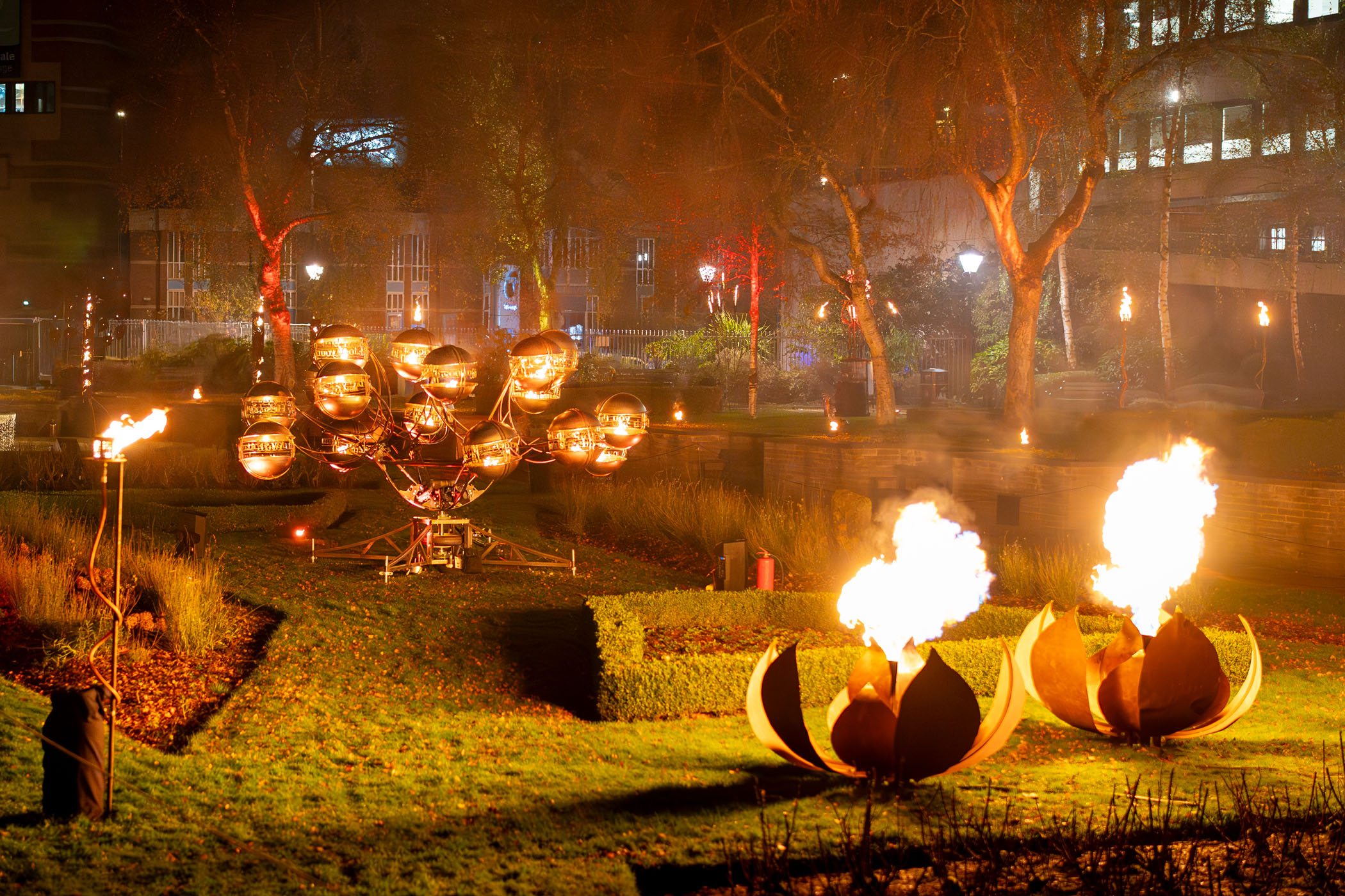 Giant metal lotus flowers breathing fire, and another fire sculpture behind it with orbs that circle. 