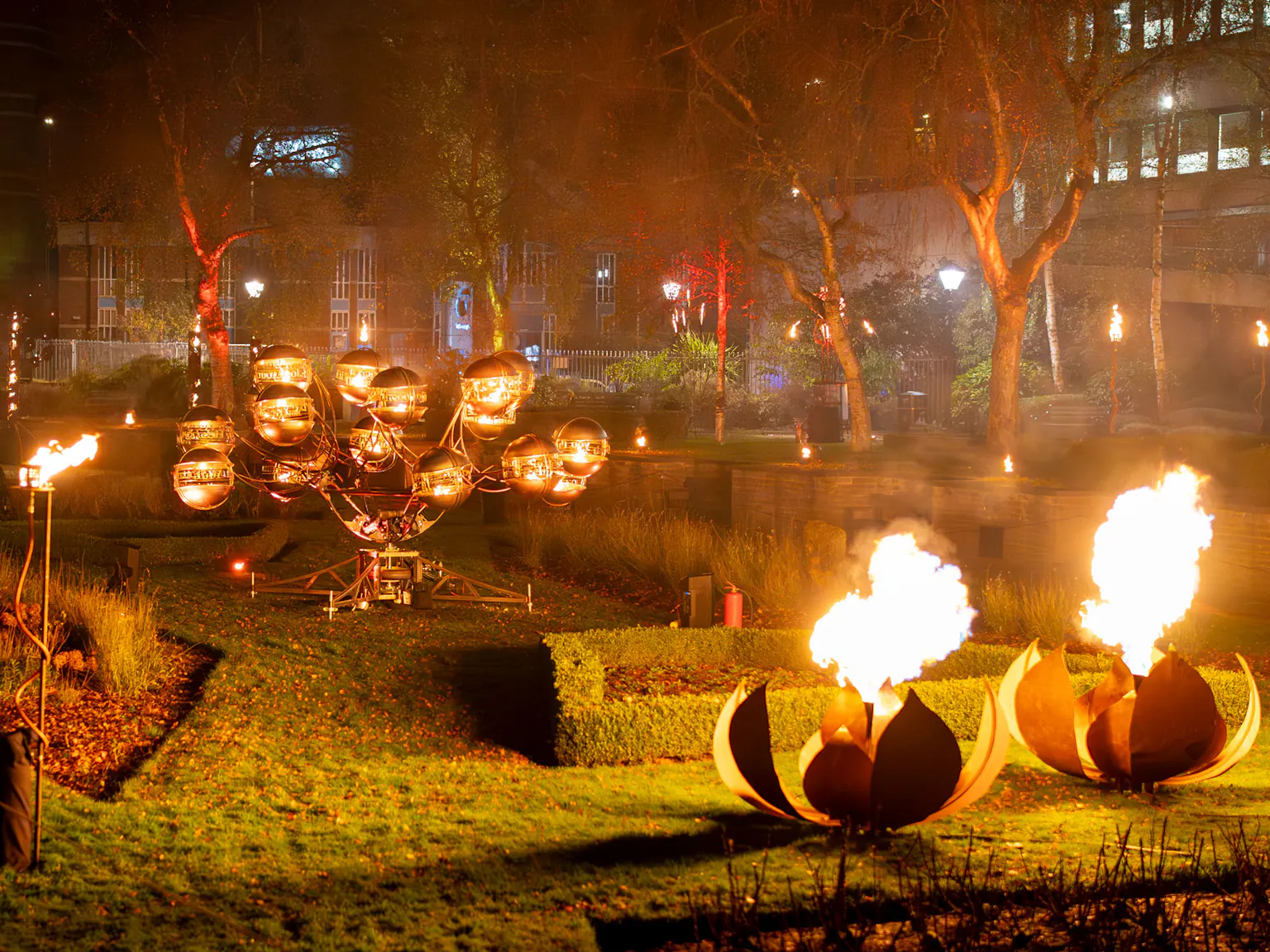 Giant metal lotus flowers breathing fire, and another fire sculpture behind it with orbs that circle.