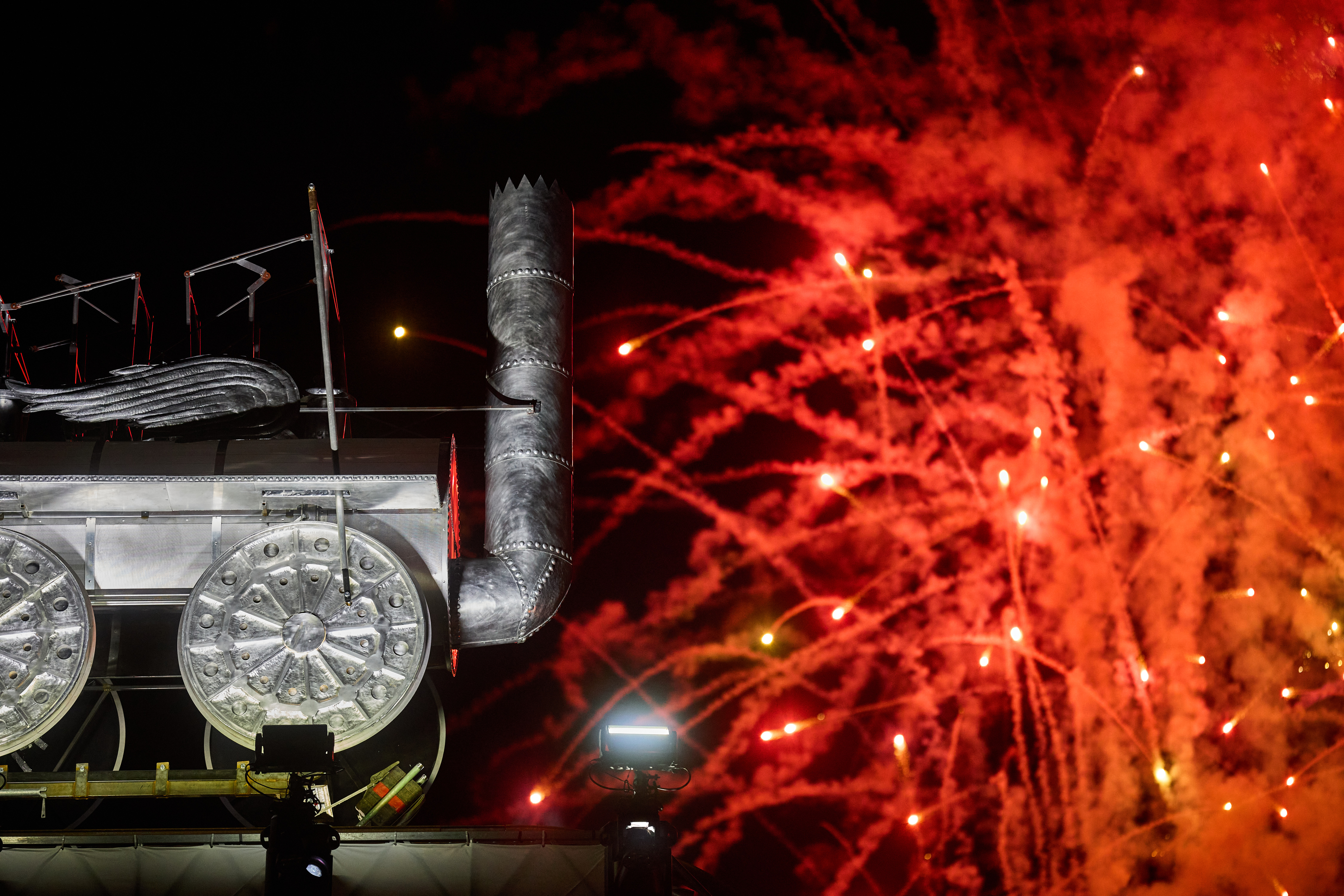A metal replica of a steam train with wings  at night with red fireworks going off behind it.