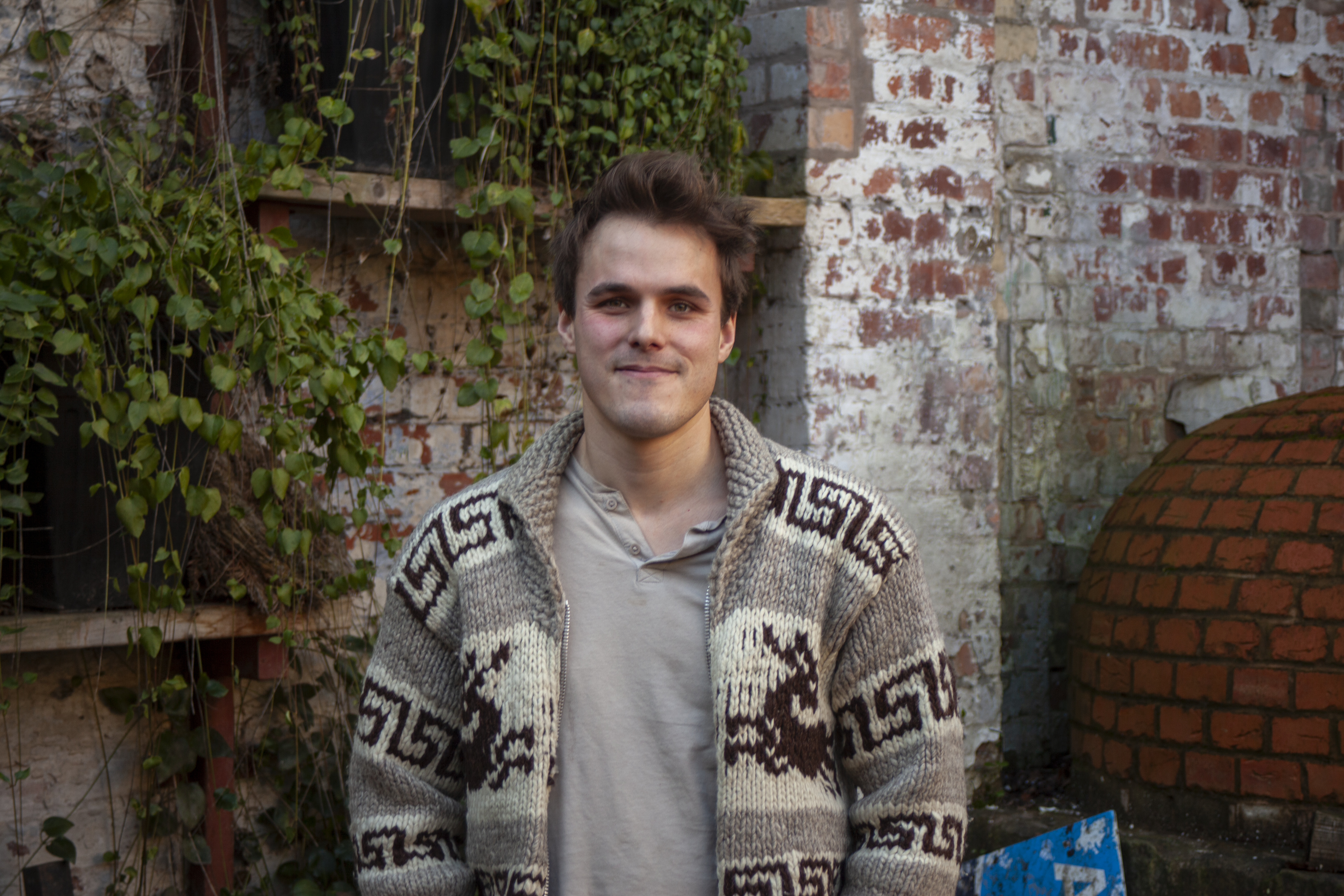 A person standing in front of greenery, he has brown hair and a grey cardigan on.