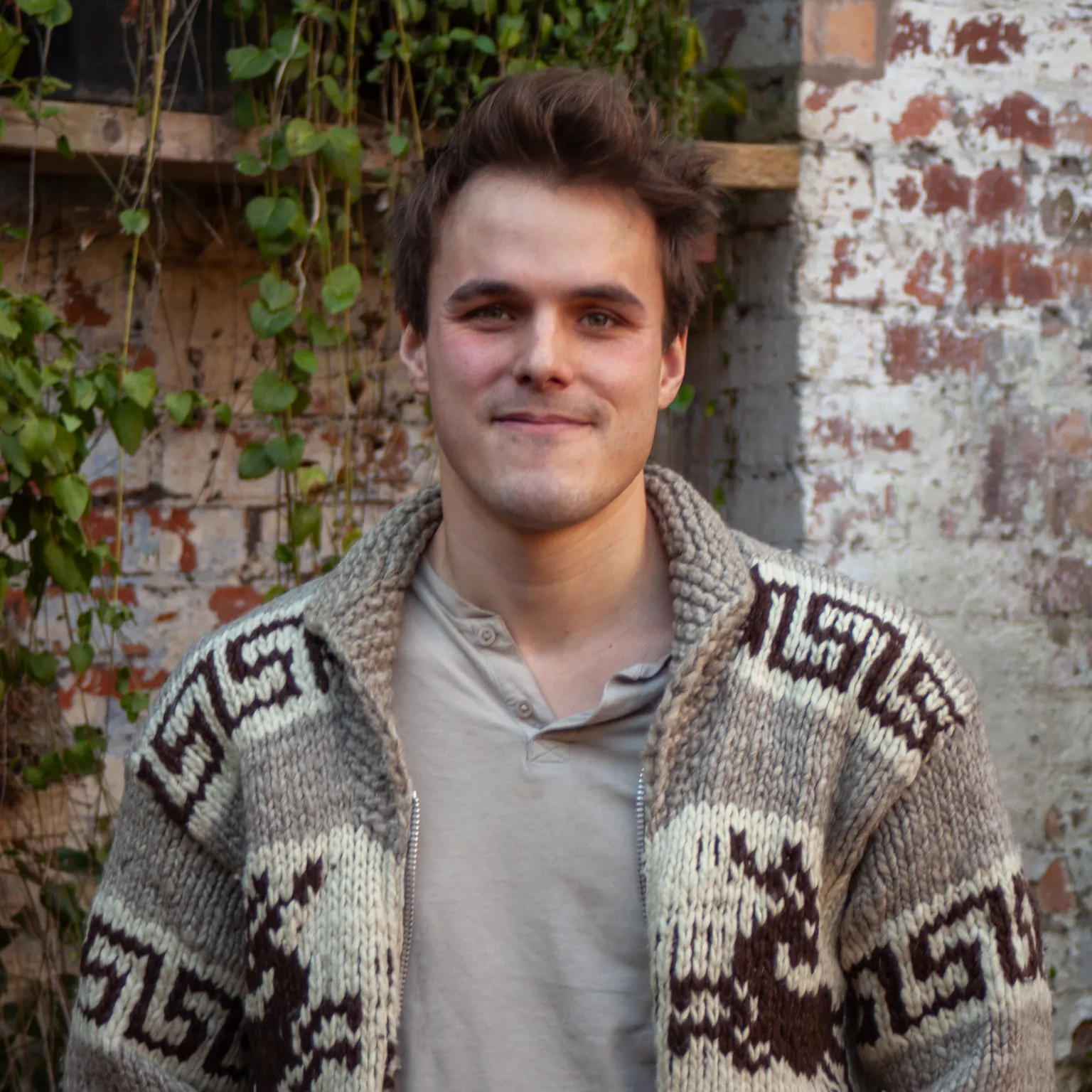 A person standing in front of greenery, he has brown hair and a grey cardigan on.