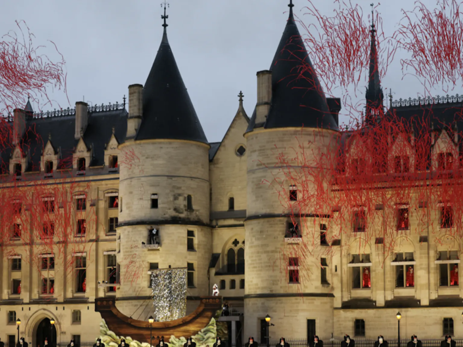 A large, majestic courthouse and prison in the Gothic Revival style featuring 2 large round turrets at the forefront and windows of various sizes set against a grey afternoon sky. Actors dressed in red gowns perform from the windows above, while actors in black line the street below. Red streamers are being fired from the ground.