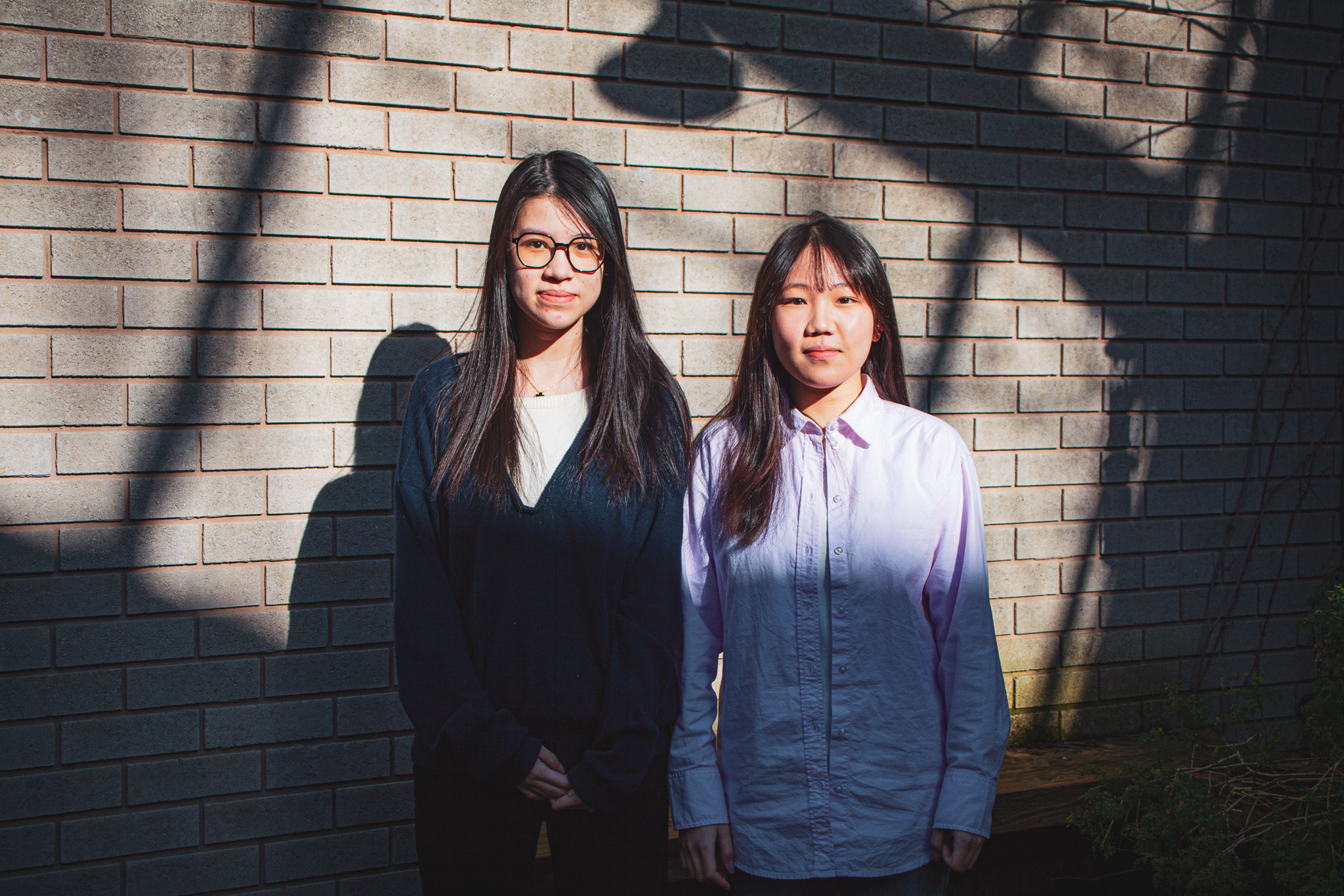 Two people with long brown hair, standing in front of a brink building with the light reflecting on them.