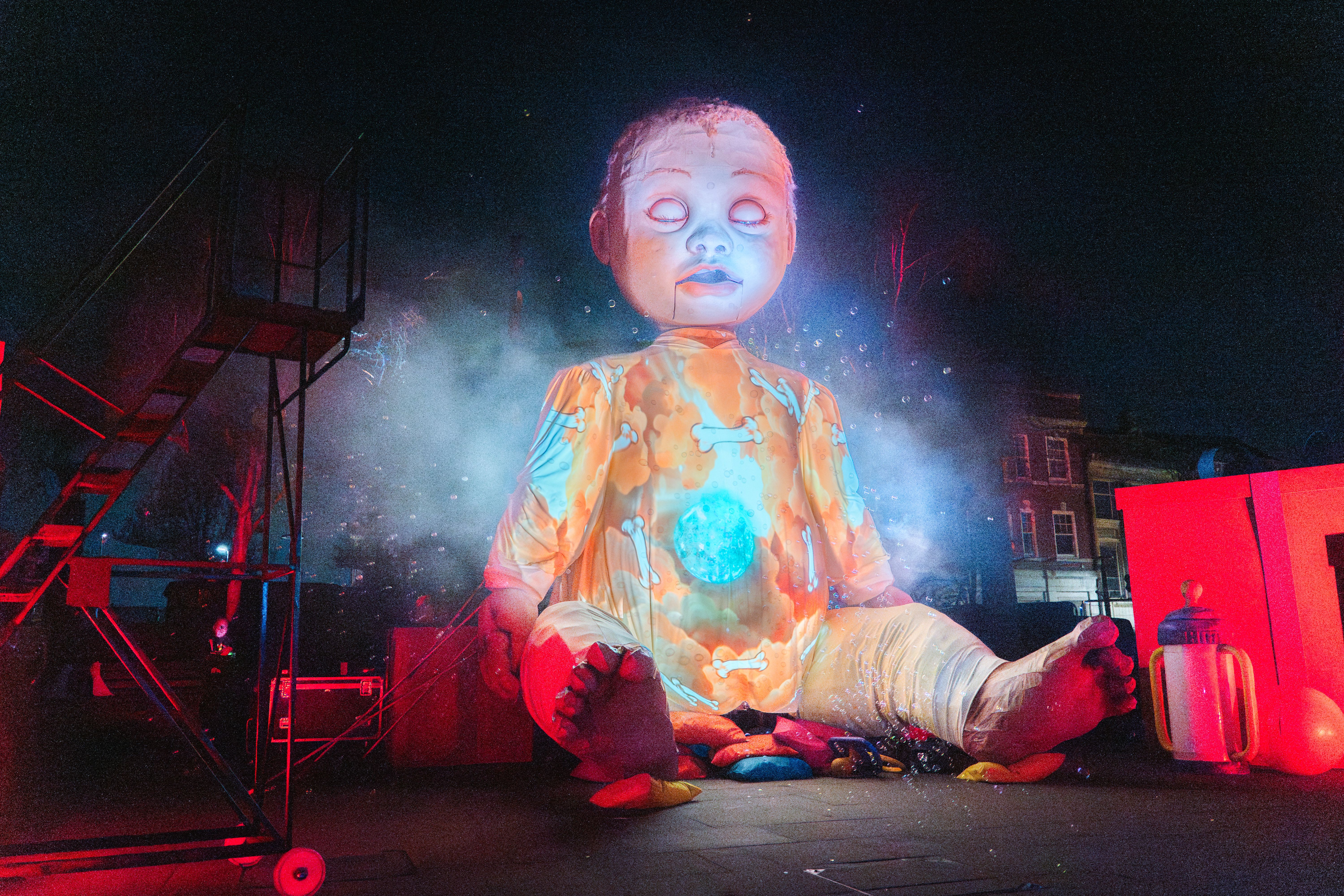 Giant illuminated baby doll sitting cross-legged outdoors at night with colourful lights and smoke around it.