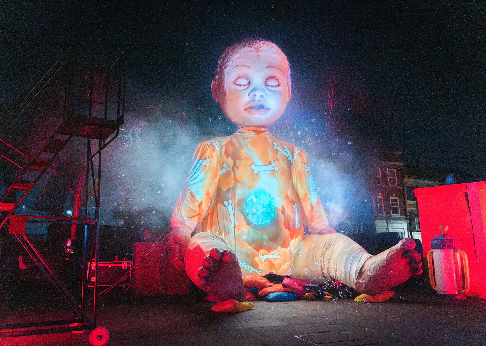 Giant illuminated baby doll sitting cross-legged outdoors at night with colourful lights and smoke around it.
