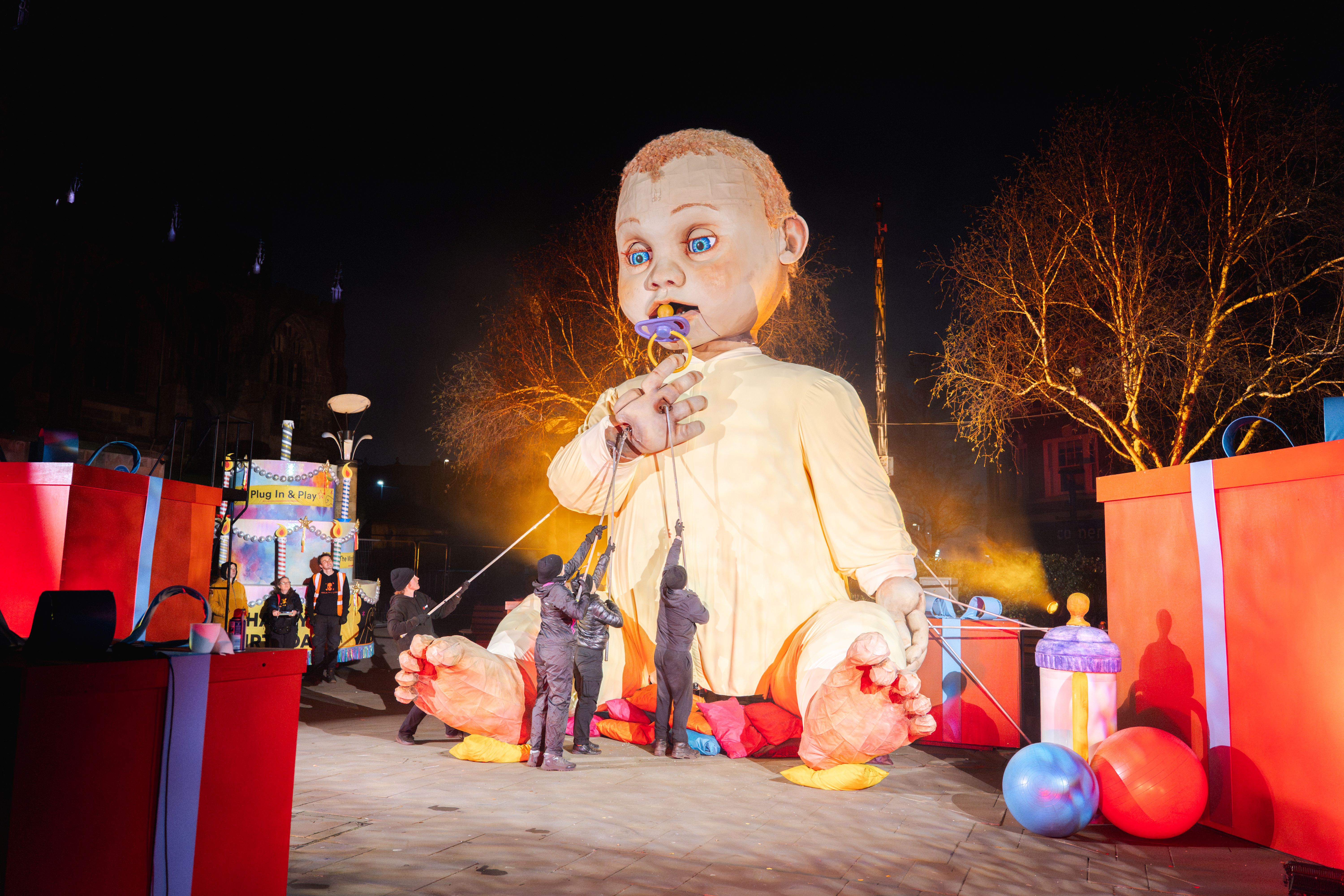 Giant puppet of a baby controlled by puppeteers during a nighttime outdoor event with large gift boxes and balloons nearby.