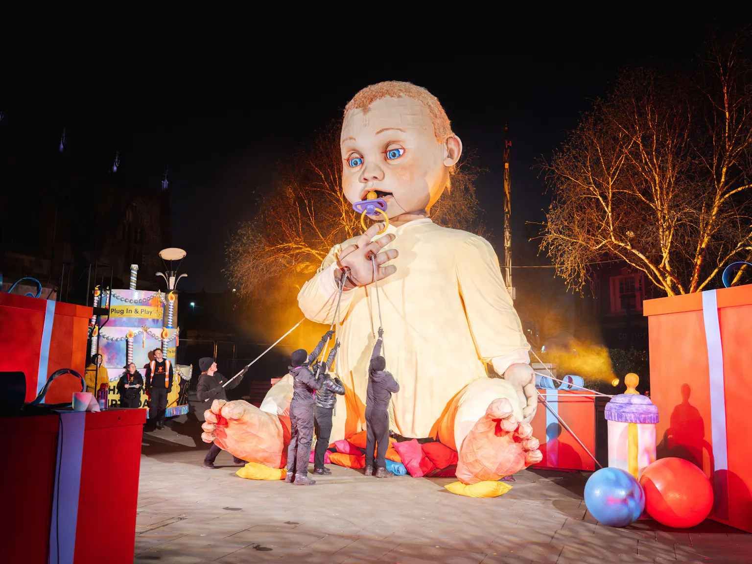 Giant puppet of a baby controlled by puppeteers during a nighttime outdoor event with large gift boxes and balloons nearby.