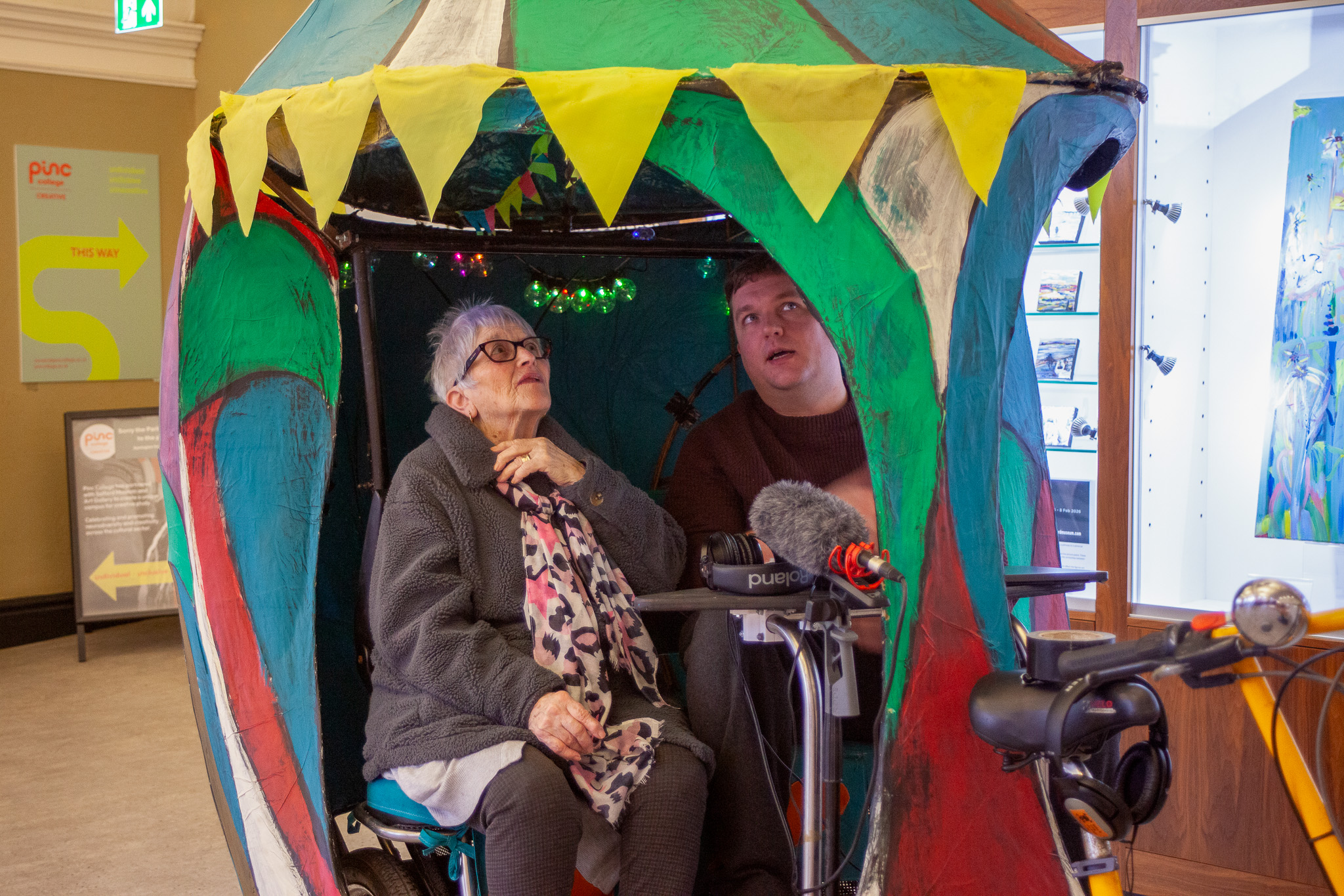 person sitting inside a colourful structure with another person standing nearby wearing headphones and operating audio equipment.