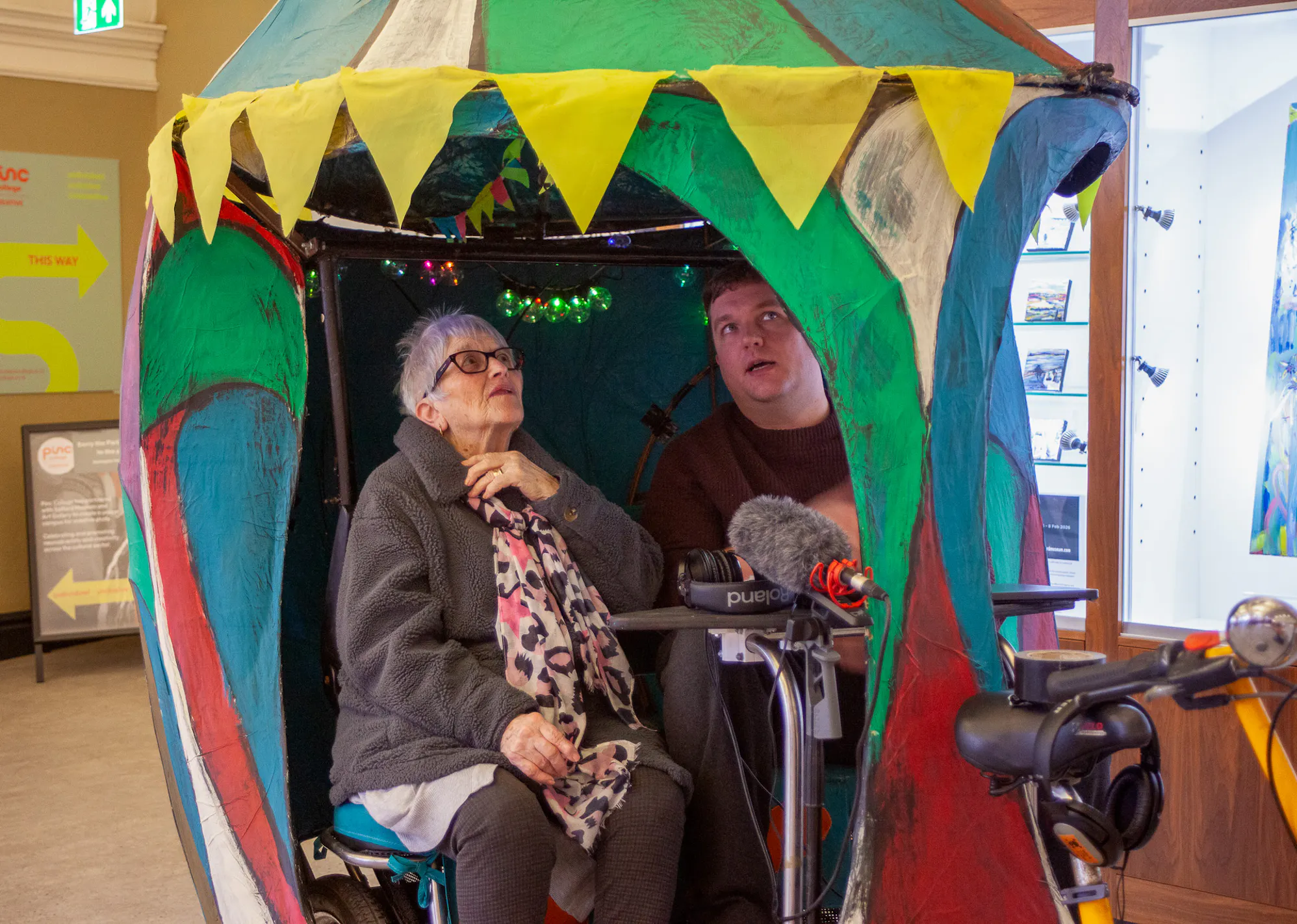 person sitting inside a colourful structure with another person standing nearby wearing headphones and operating audio equipment.