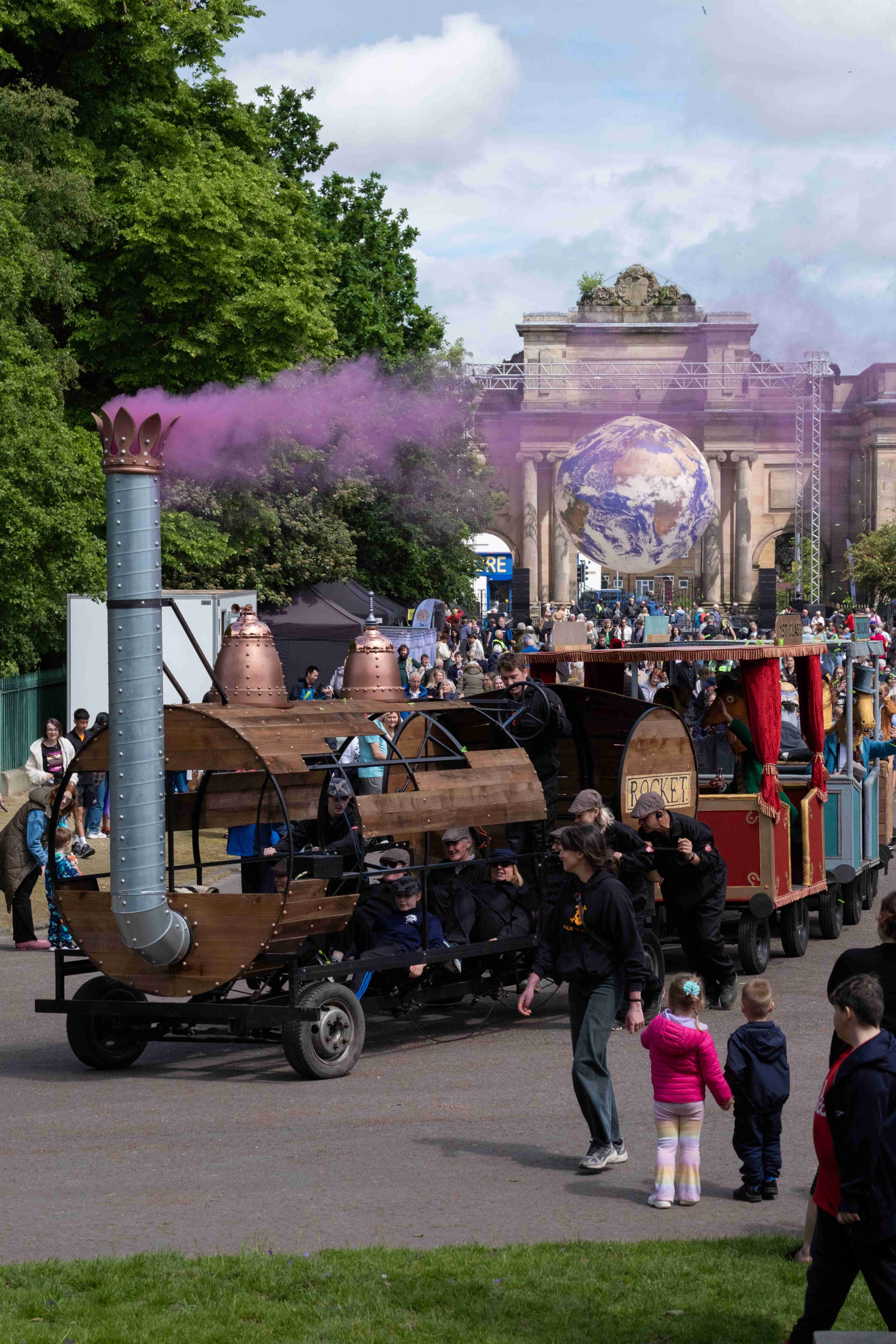 A train float with three carriages carrying pantomime horses in park as crowd look on with pink smoke.
