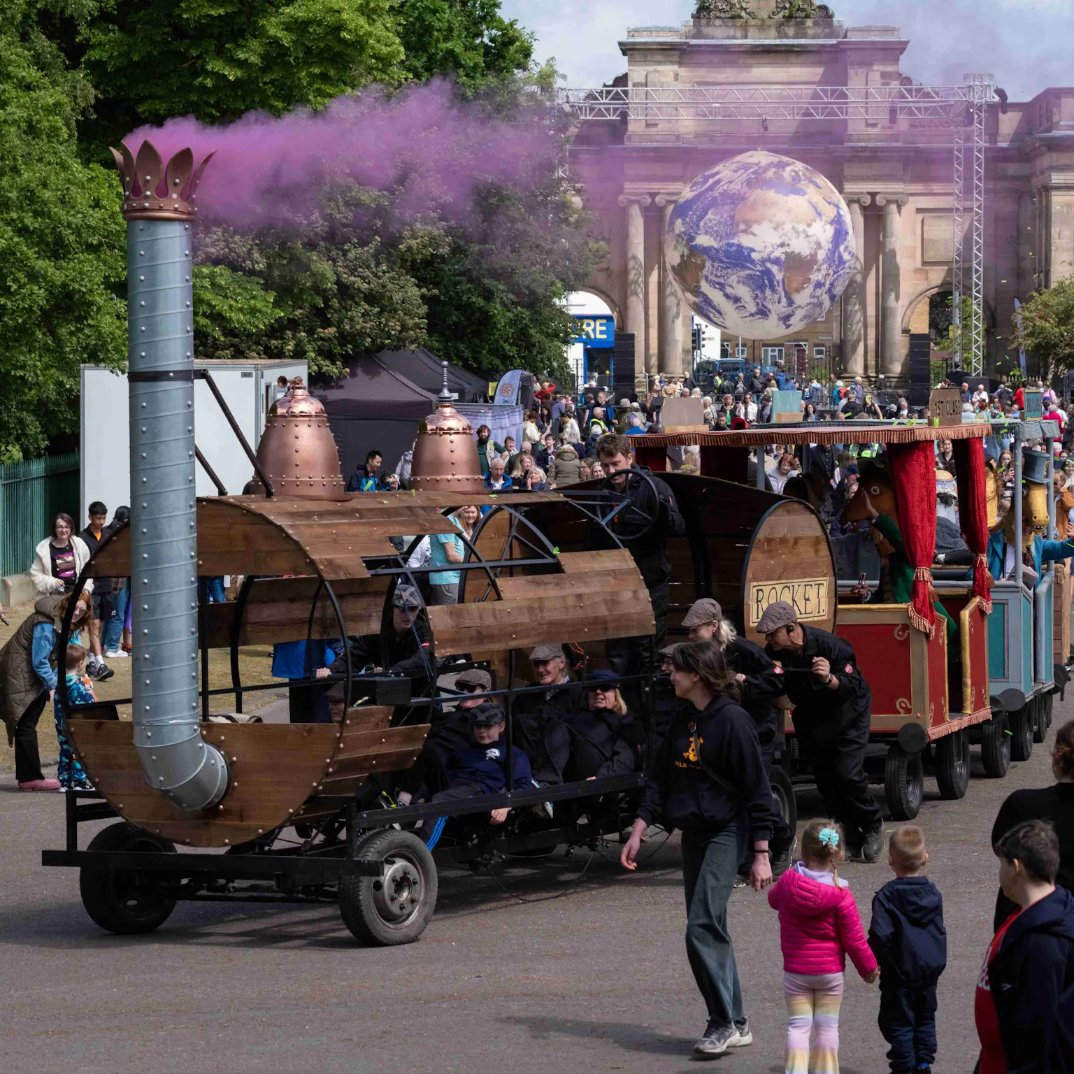 A train float with three carriages carrying pantomime horses in park as crowd look on with pink smoke.