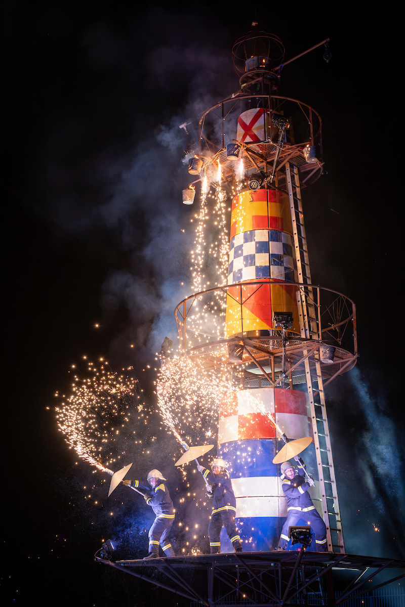 A large lighthouse structure with three lighthouse keeper performers on the structure with pyrotechnics