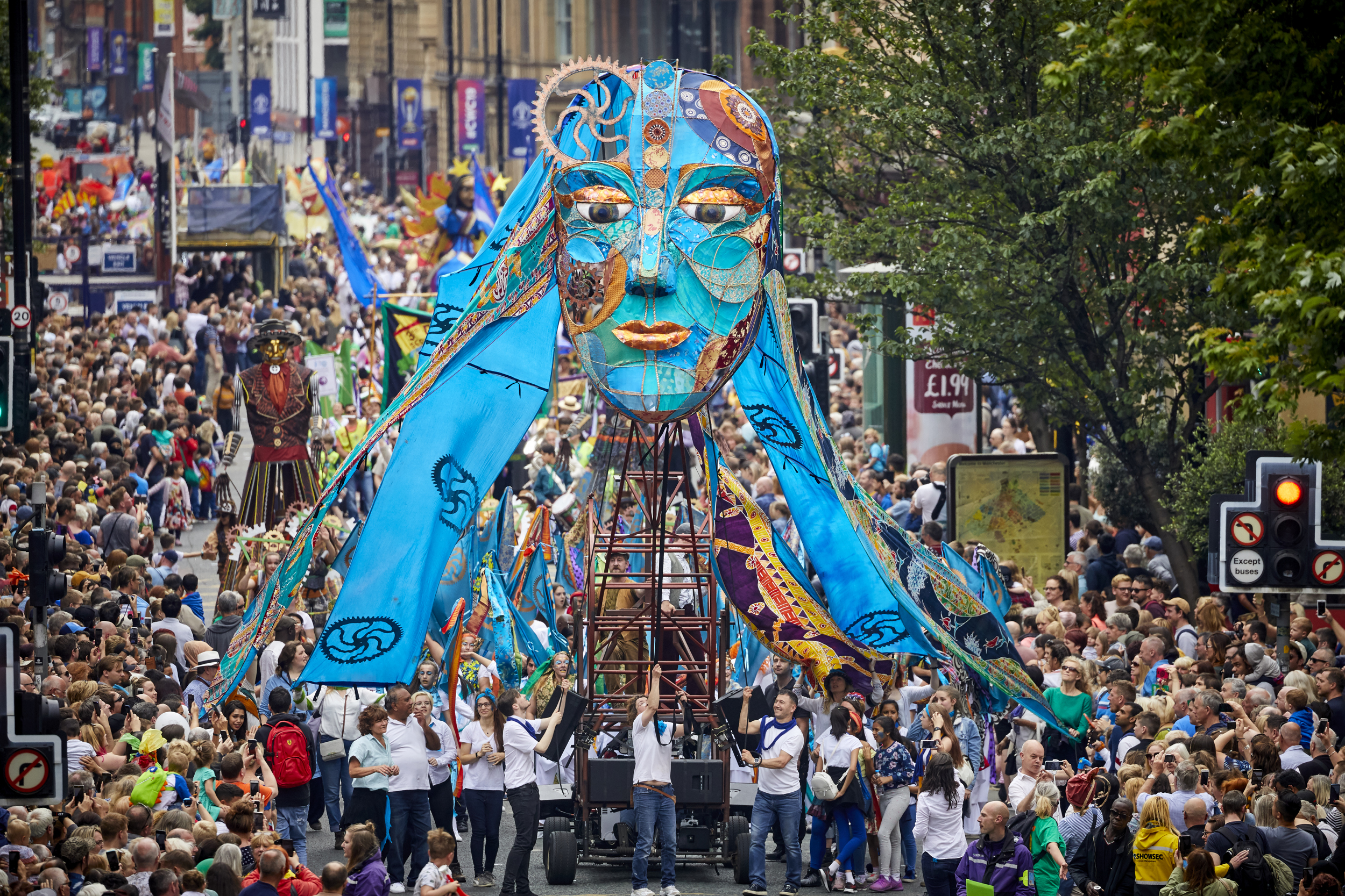 A large blue face being paraded above the crowd along a busy road