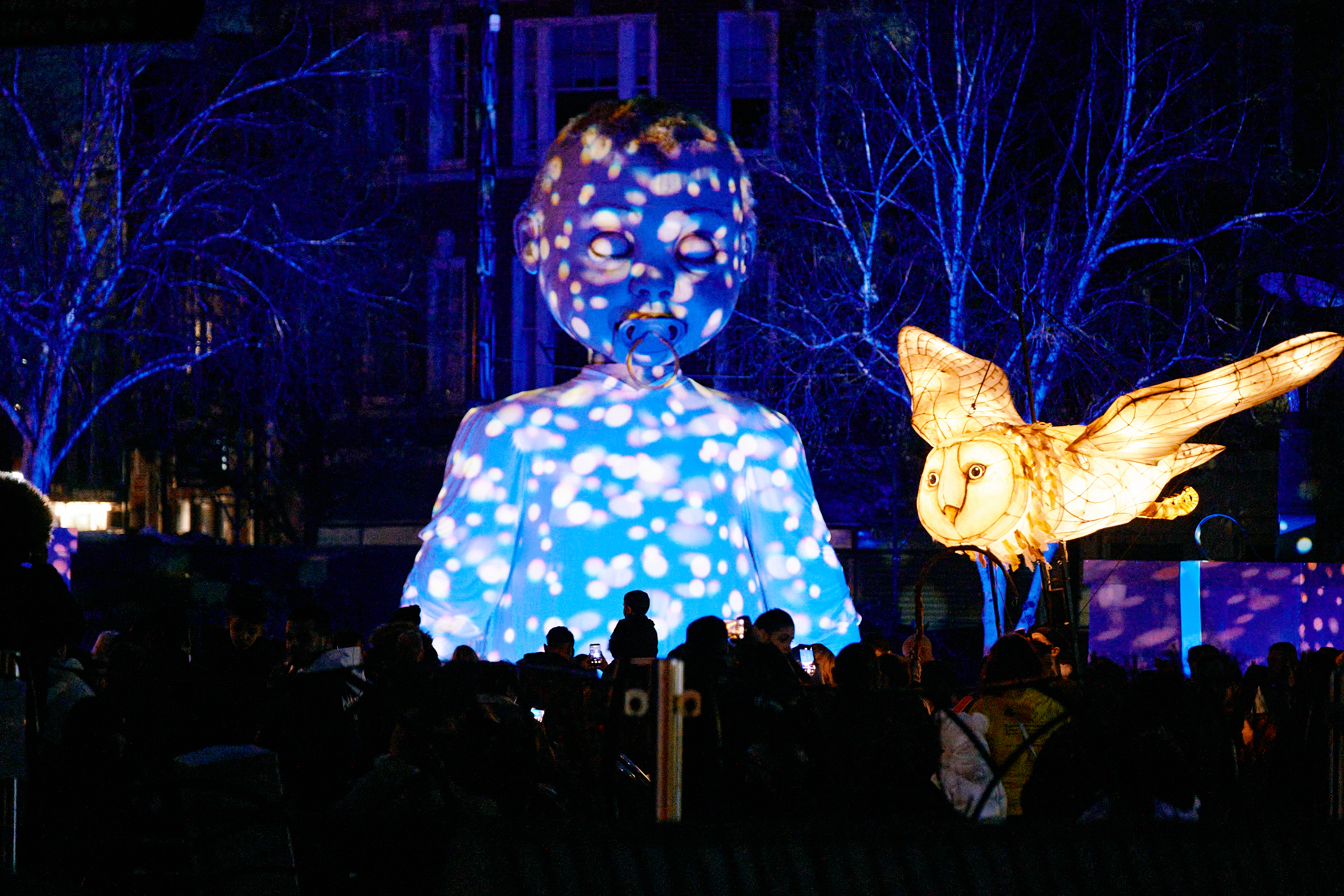 A baby puppet is lit up with projections as a white owl puppet illuminates the sky.