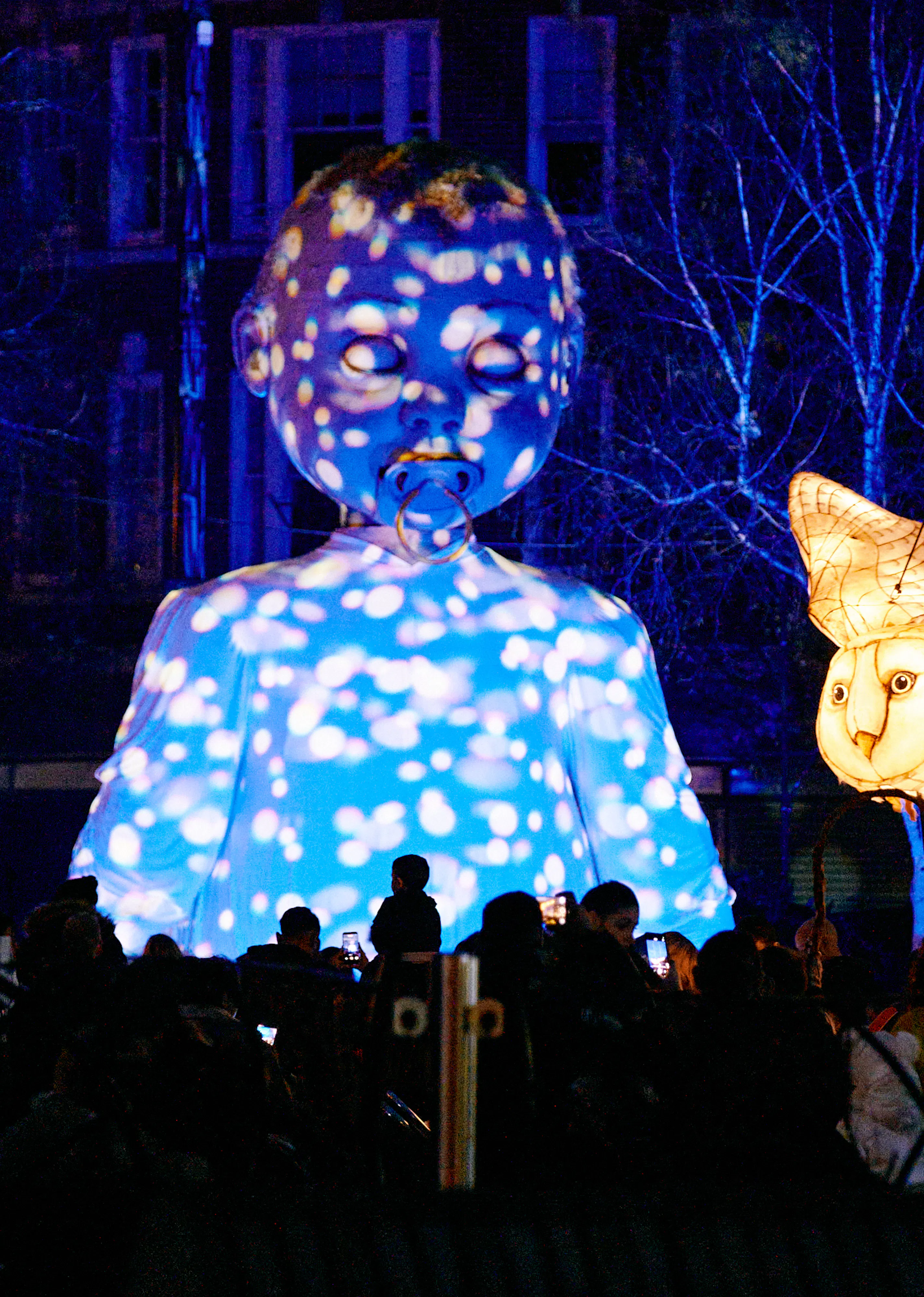 A baby puppet is lit up with projections as a white owl puppet illuminates the sky.