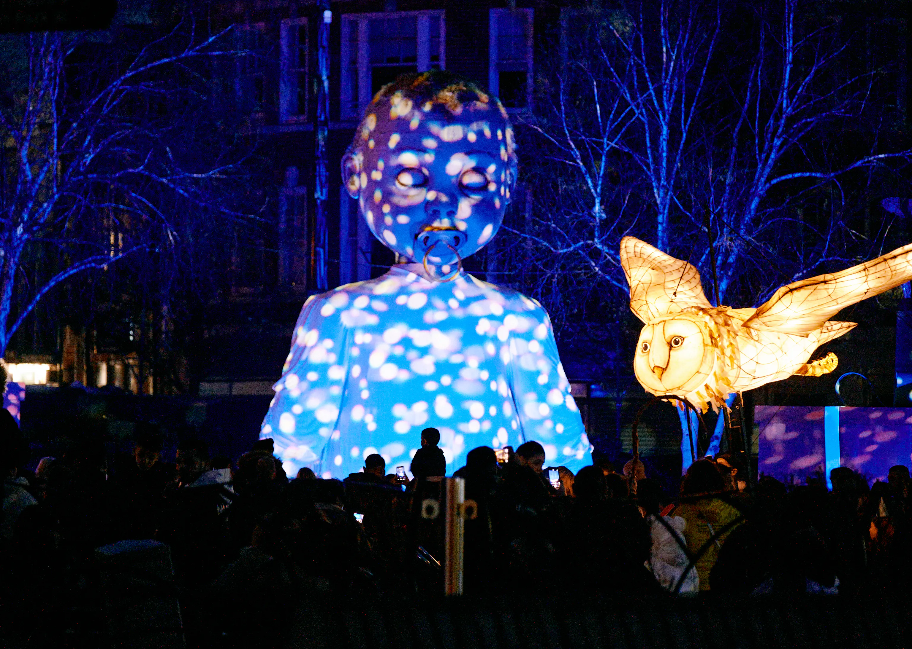 A baby puppet is lit up with projections as a white owl puppet illuminates the sky.