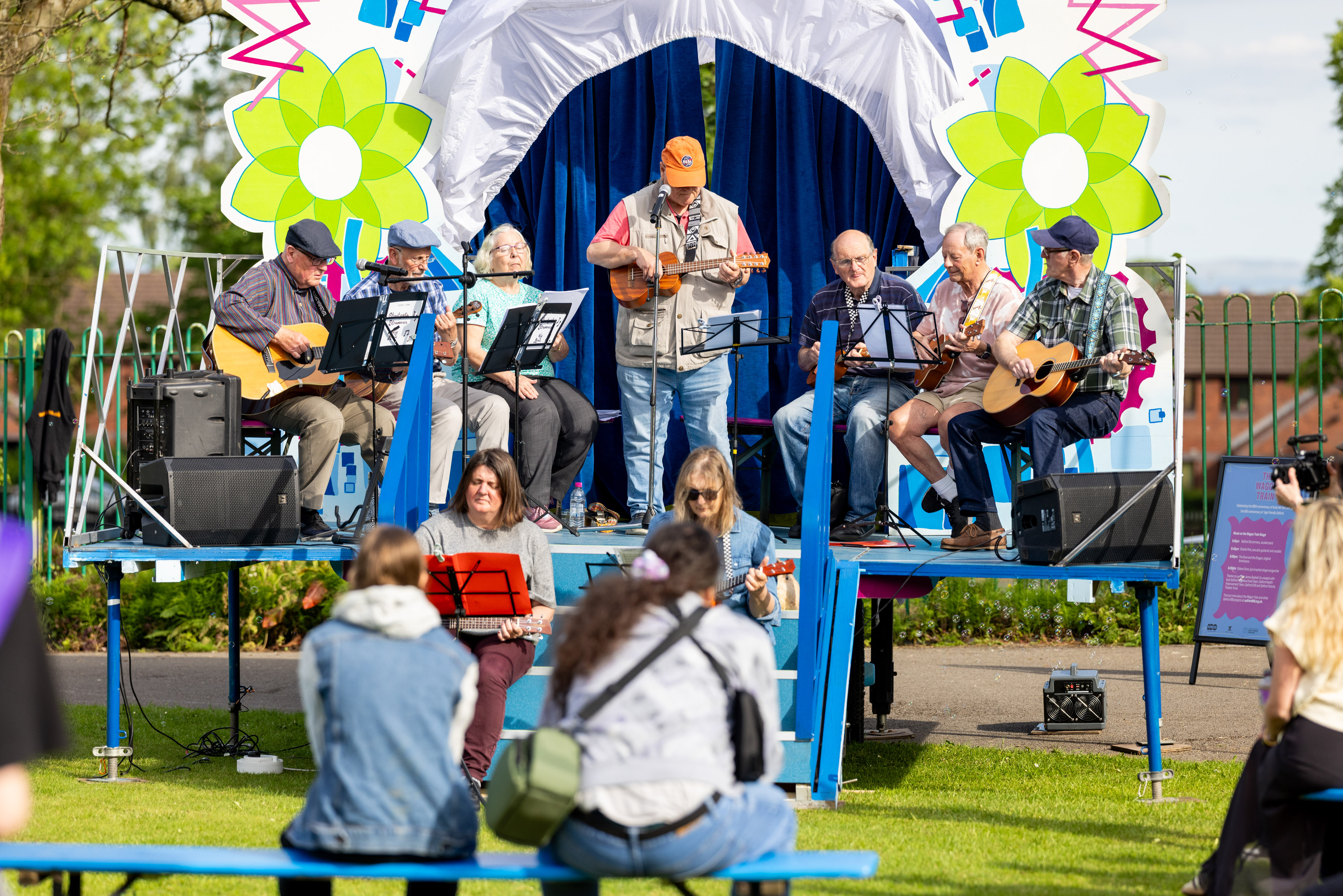 Group of elderly musicians playing guitars and ukuleles on a decorated outdoor stage during a sunny day event.