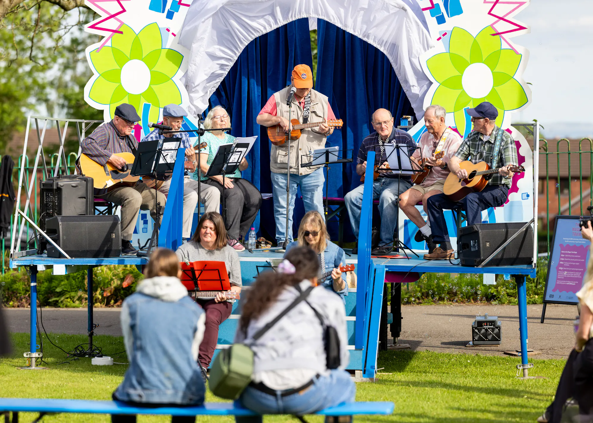 Group of elderly musicians playing guitars and ukuleles on a decorated outdoor stage during a sunny day event.