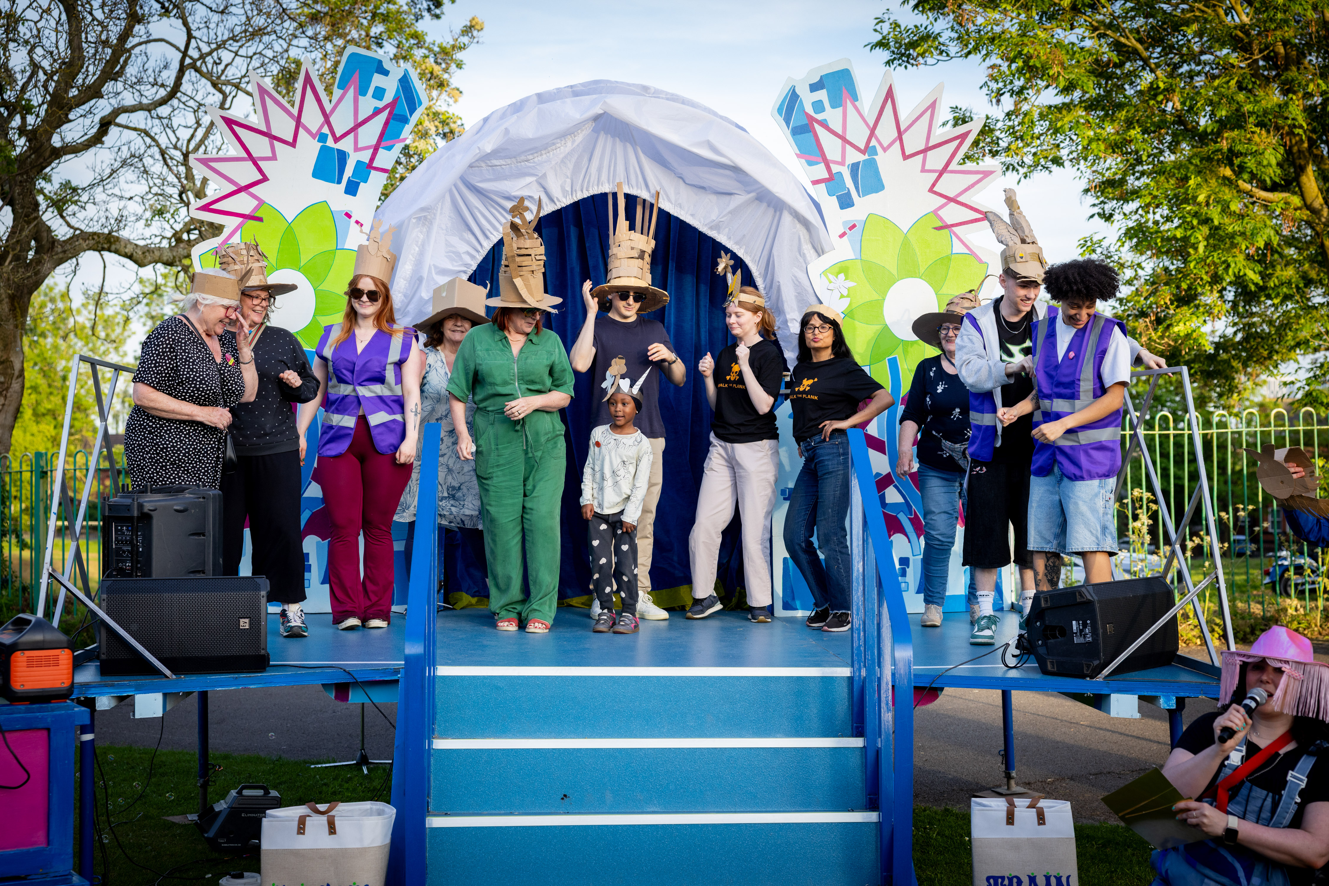 Group of people on a decorated outdoor stage wearing colorful costumes and hats, engaging in a lively performance or celebration.