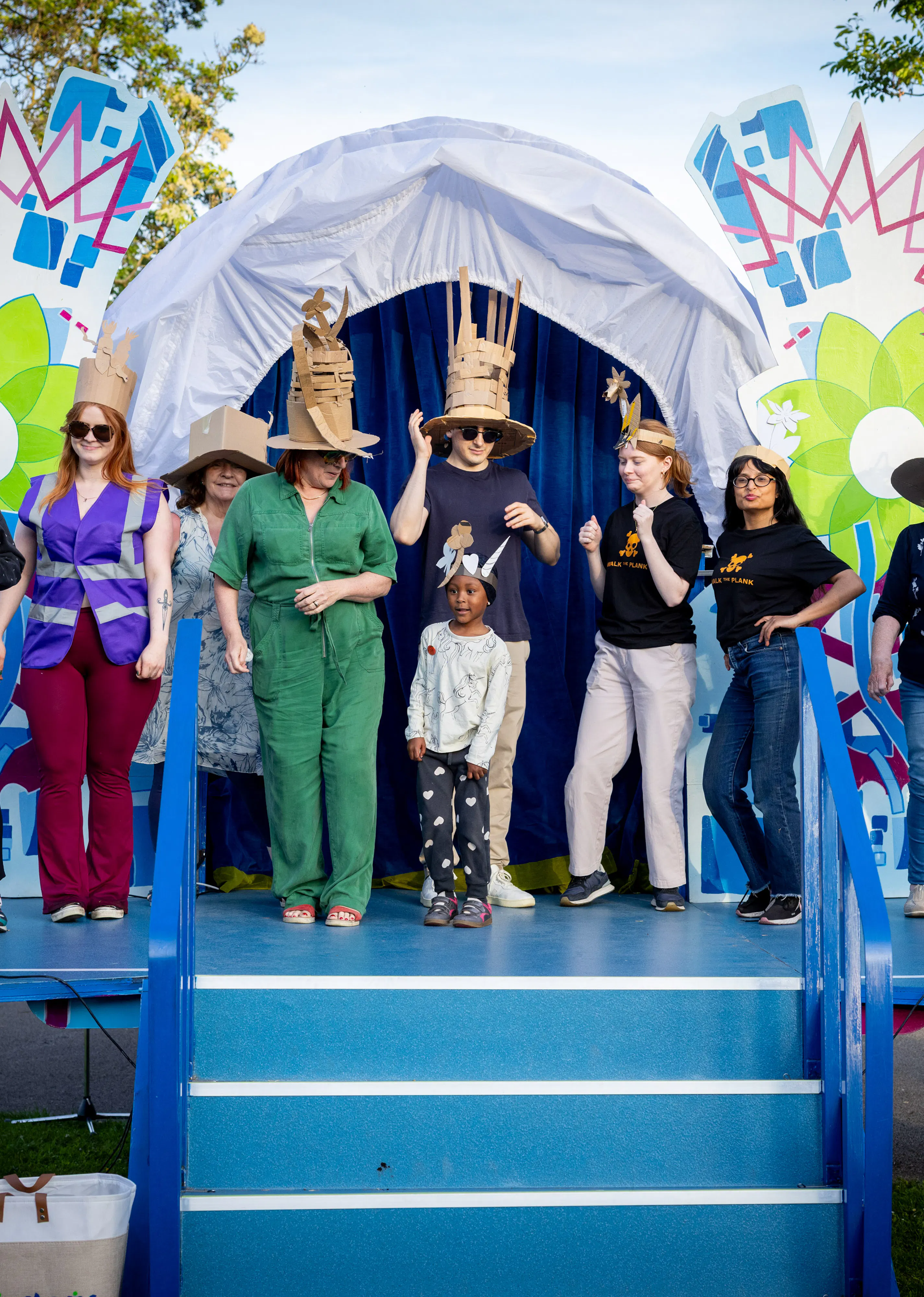 Group of people on a decorated outdoor stage wearing colorful costumes and hats, engaging in a lively performance or celebration.