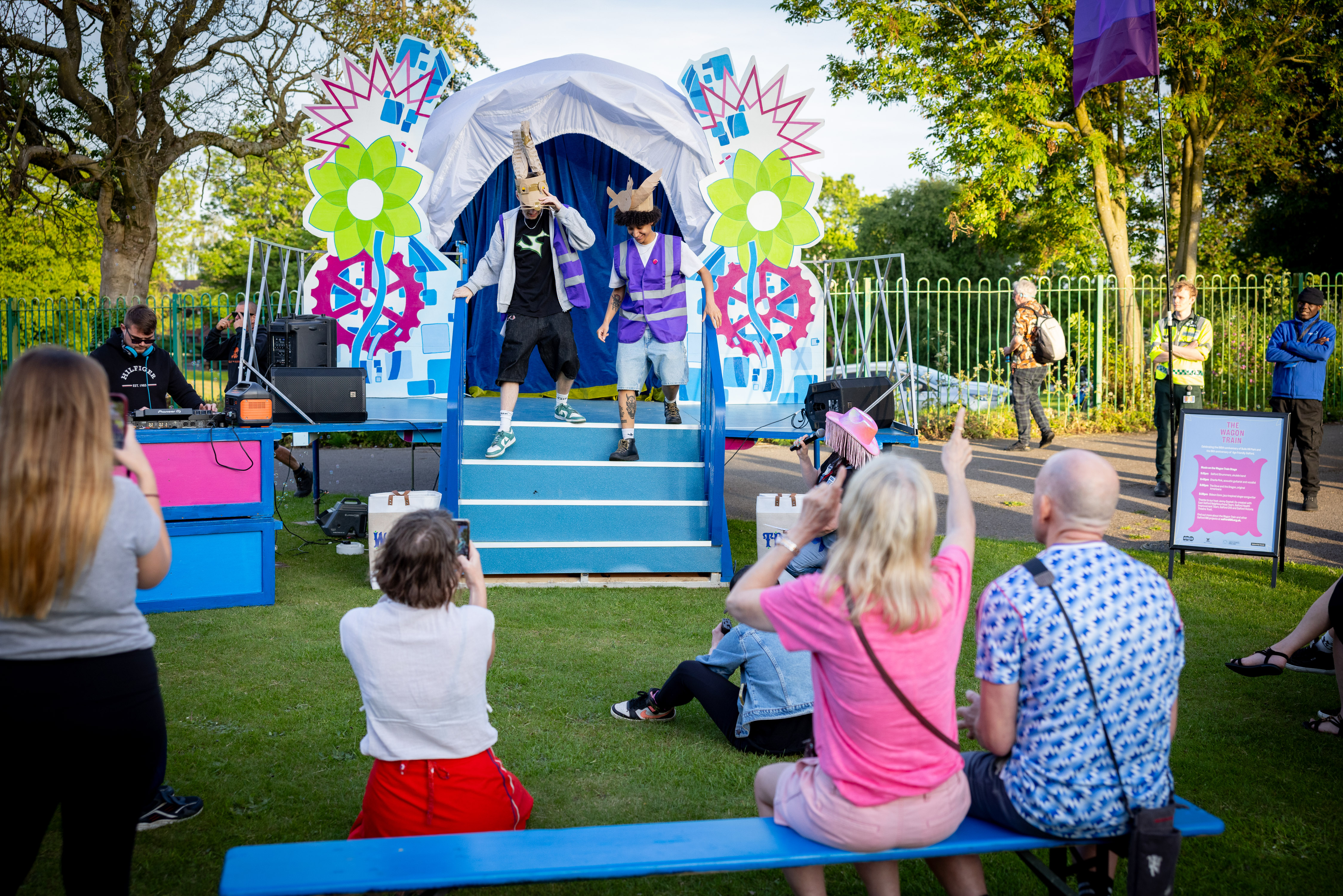 Two performers in colourful costumes entertain an outdoor audience in front of a decorated stage with bright flowers and gears.


