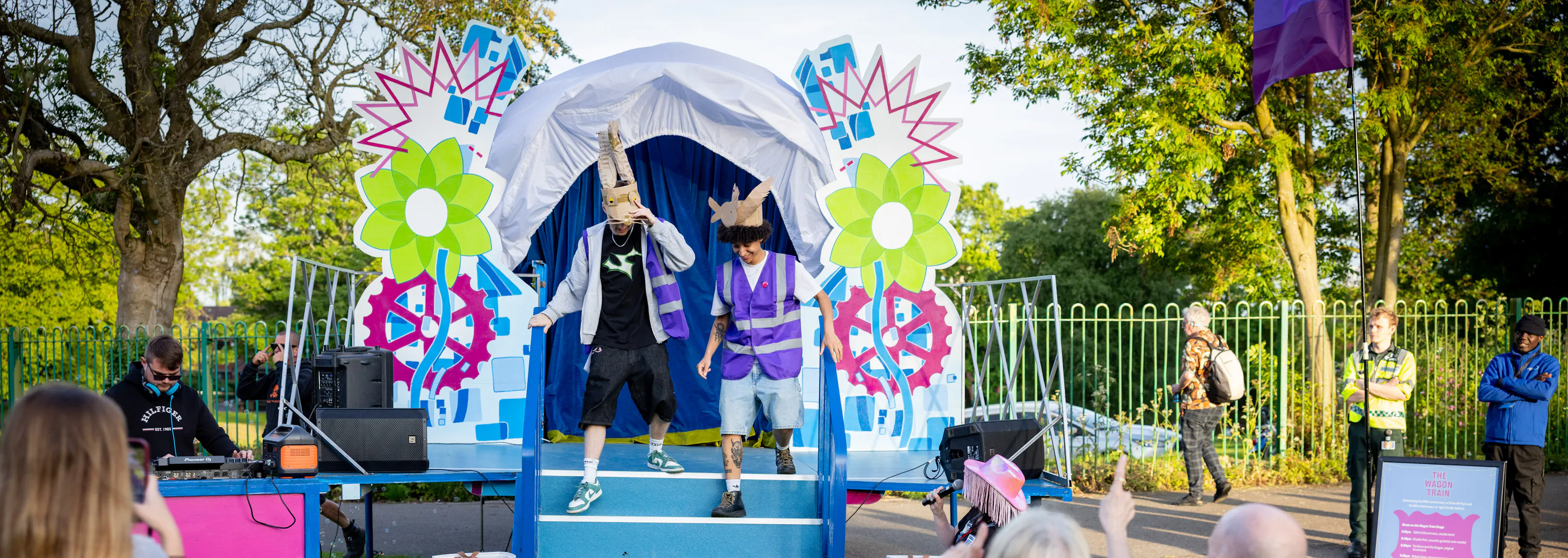 Two performers in colourful costumes entertain an outdoor audience in front of a decorated stage with bright flowers and gears.
