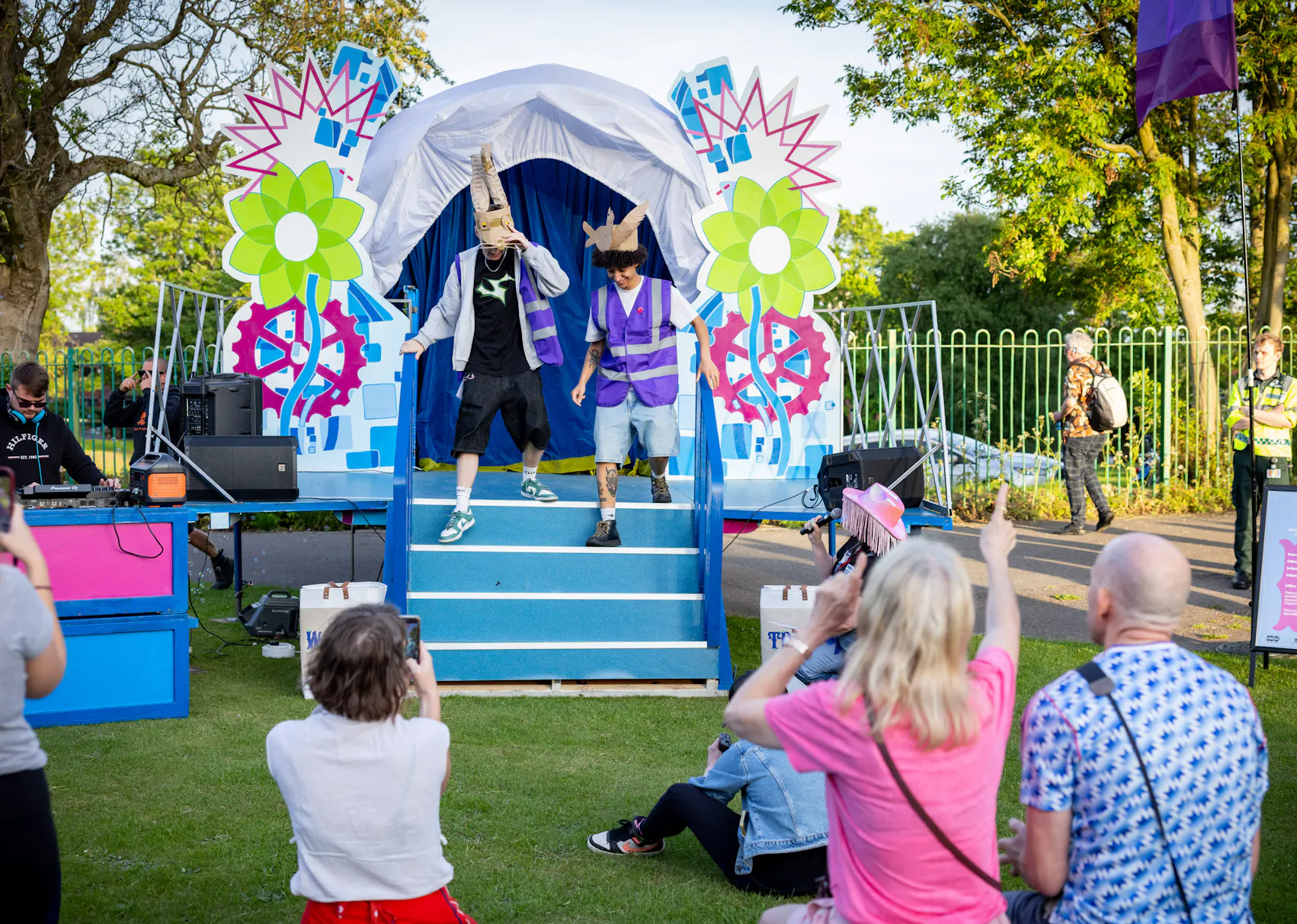 Two performers in colourful costumes entertain an outdoor audience in front of a decorated stage with bright flowers and gears.