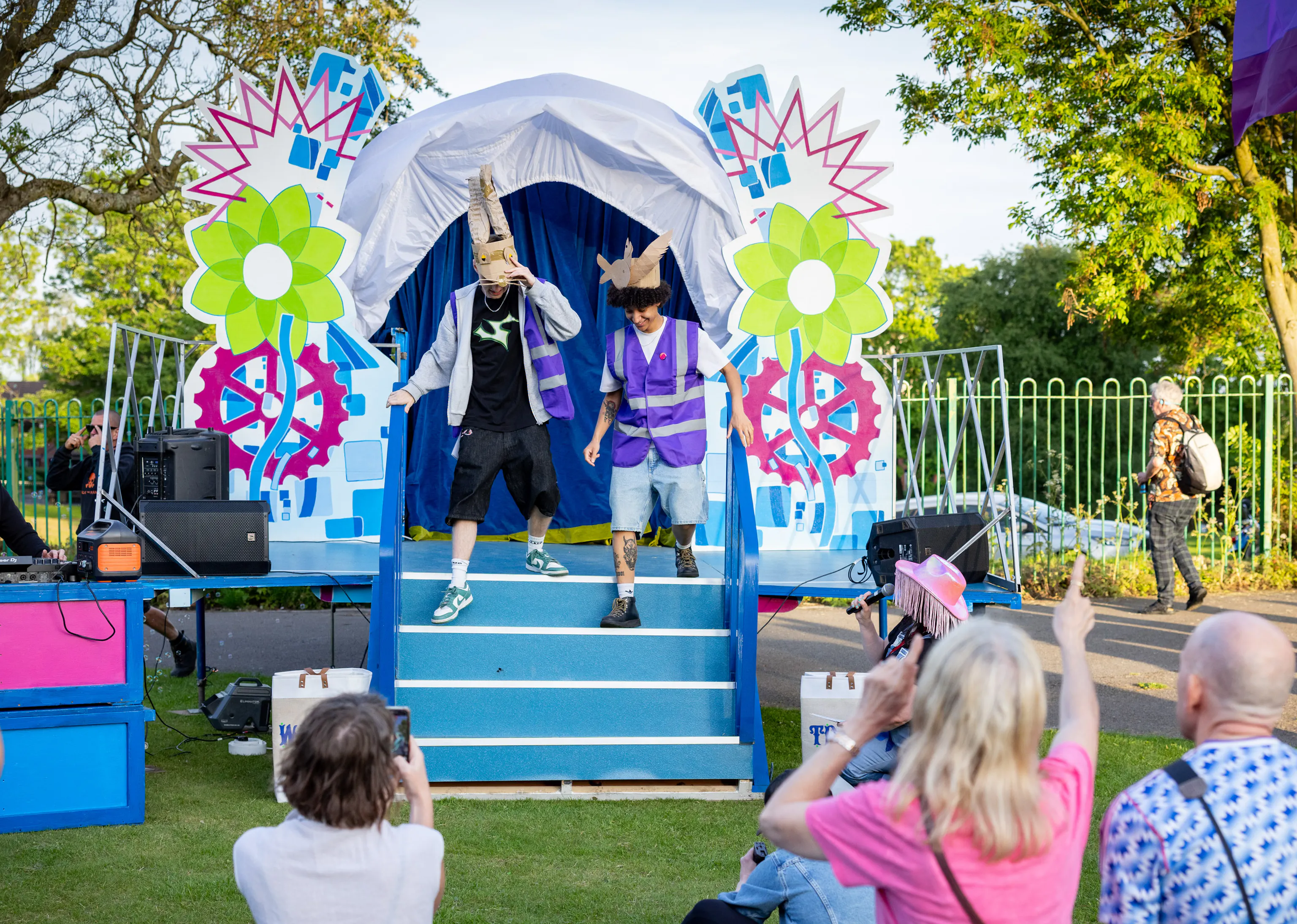 Two performers in colourful costumes entertain an outdoor audience in front of a decorated stage with bright flowers and gears.