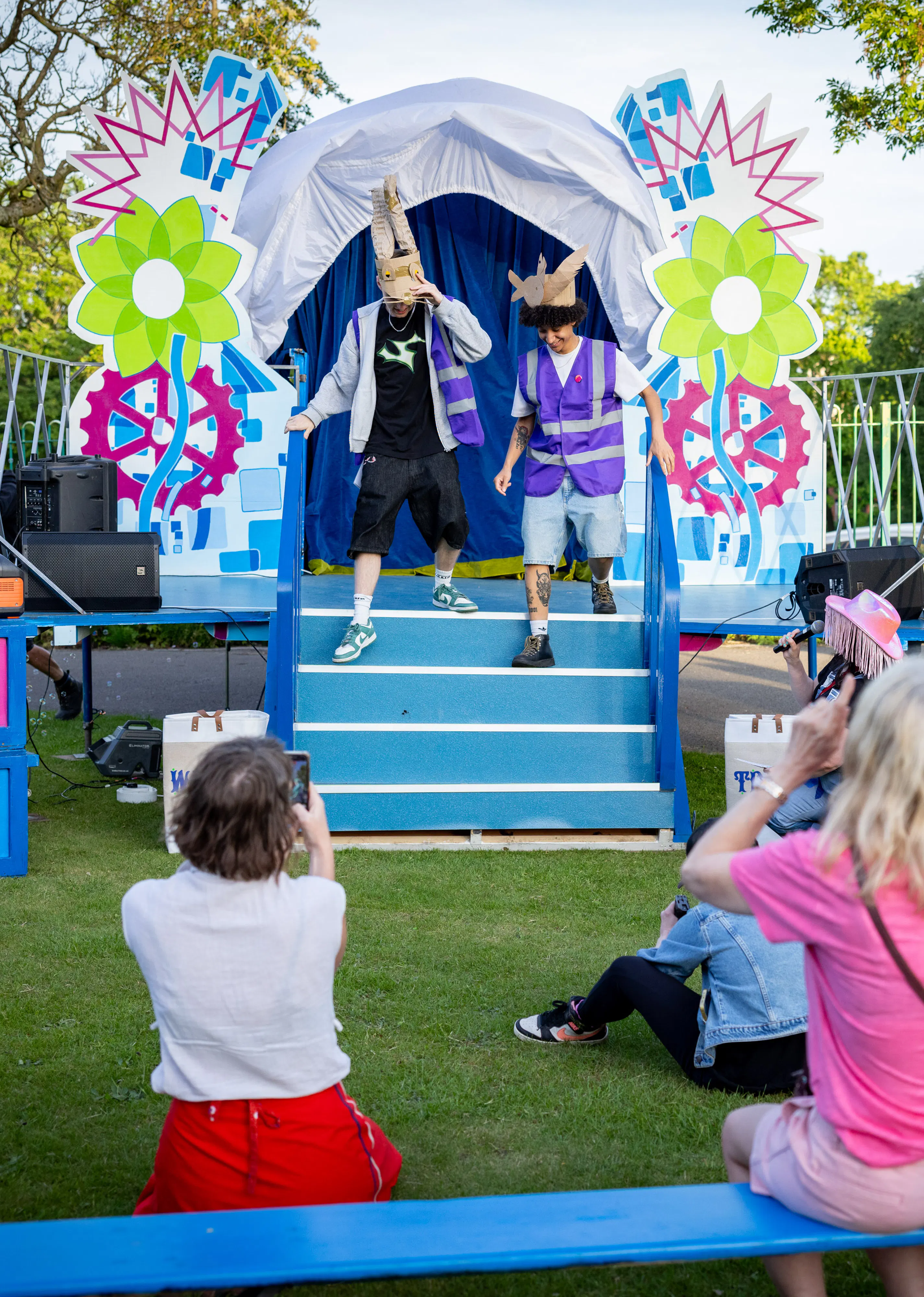 Two performers in colourful costumes entertain an outdoor audience in front of a decorated stage with bright flowers and gears.